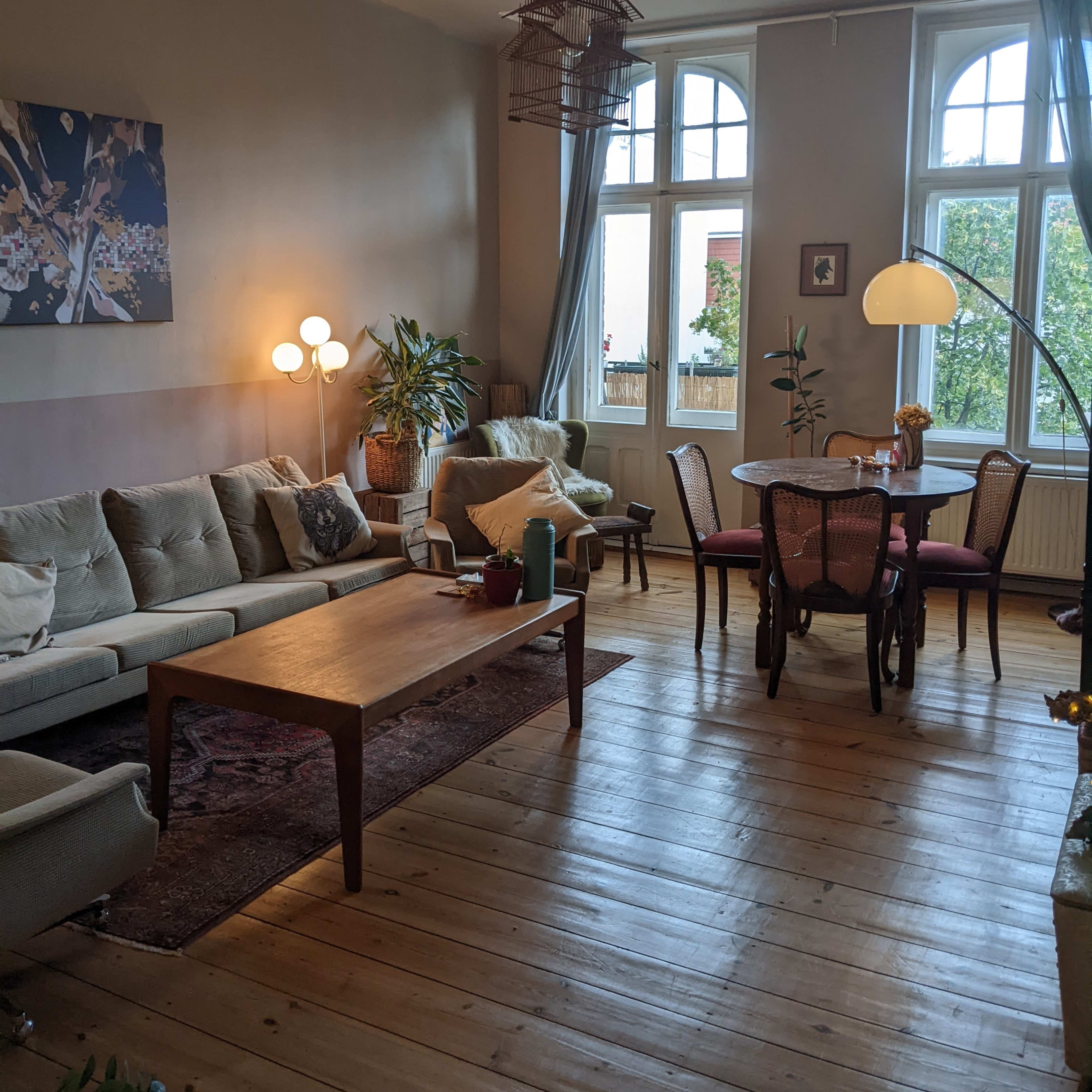A spacious living room features a light-colored sectional sofa, a wooden coffee table, a dining area with wicker chairs, and large windows allowing natural light.