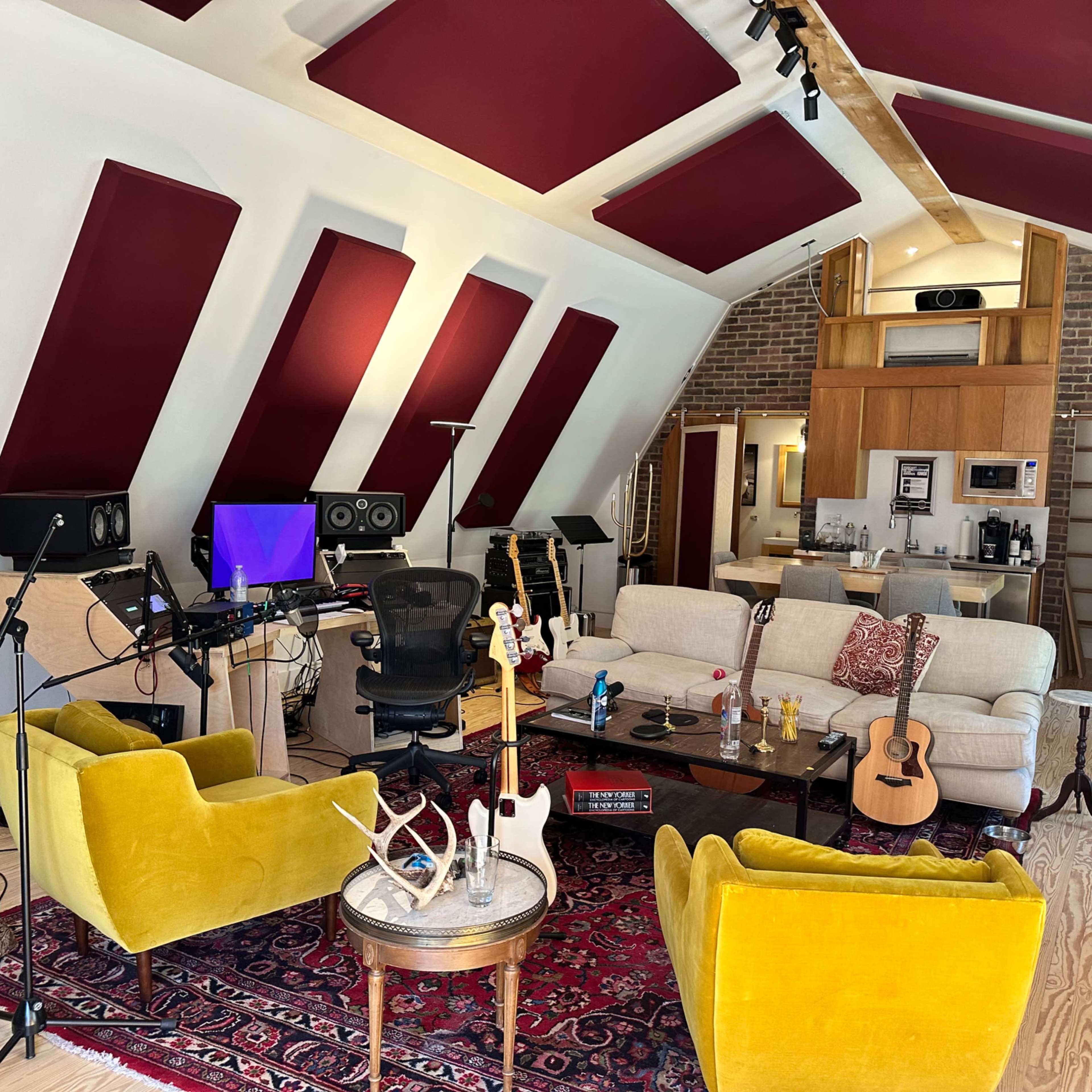 A music studio with a central seating area featuring yellow armchairs, a sofa, musical instruments, and sound equipment against a backdrop of angled walls and a kitchenette.