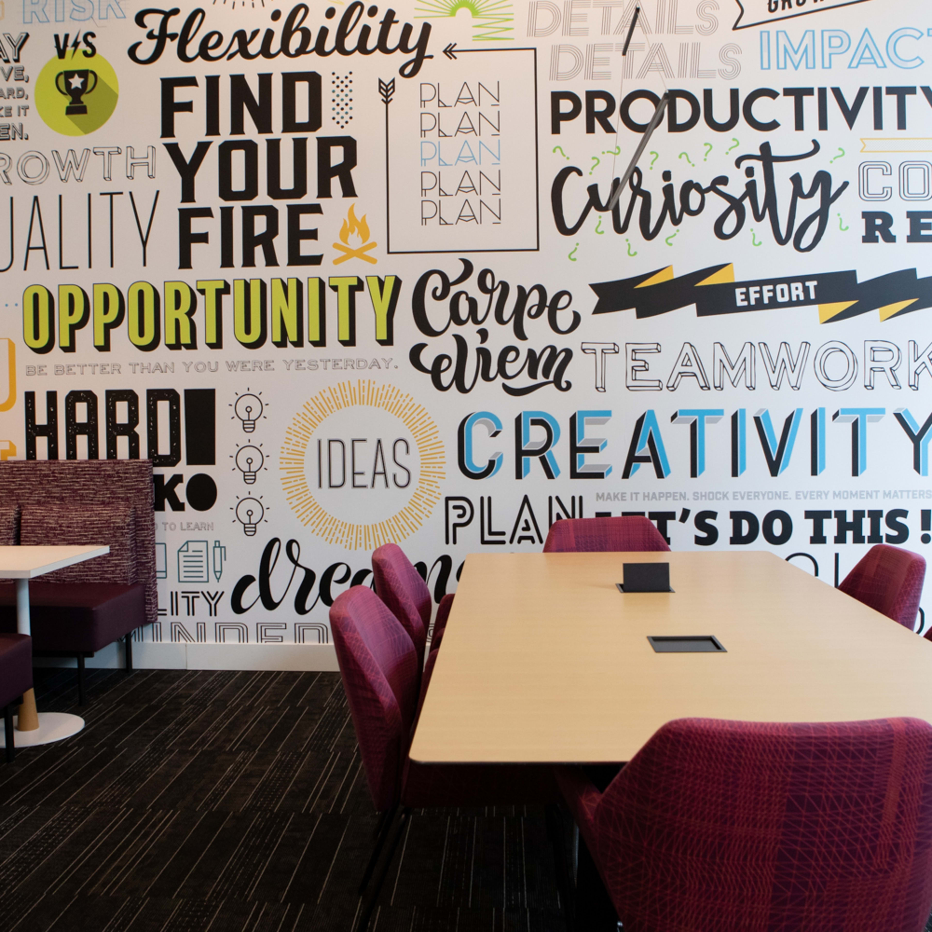 A modern meeting room with a large table surrounded by red chairs, featuring a wall covered in motivational words and phrases.