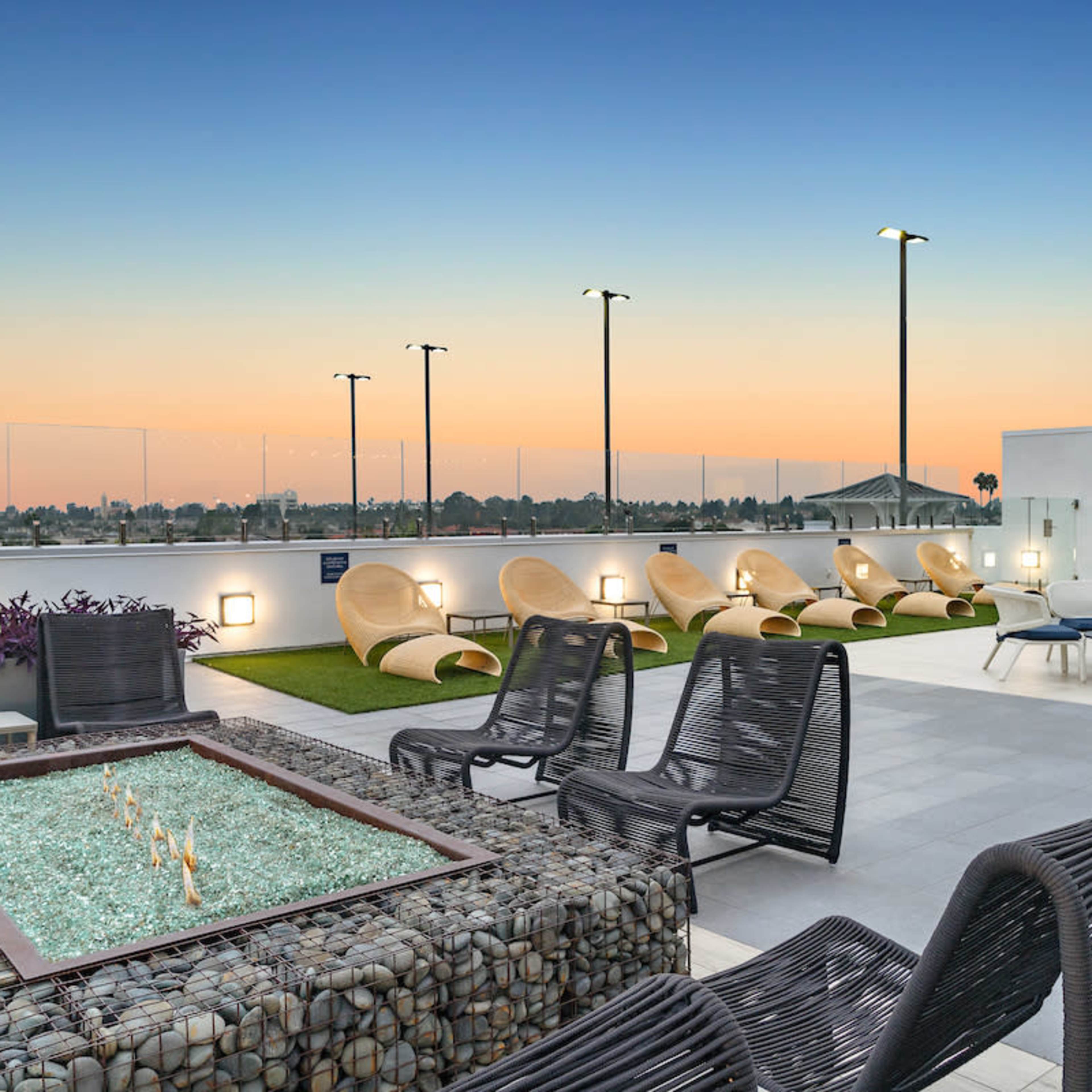 The rooftop terrace features seating areas, a fire pit, and lounge chairs against a backdrop of a sunset sky.