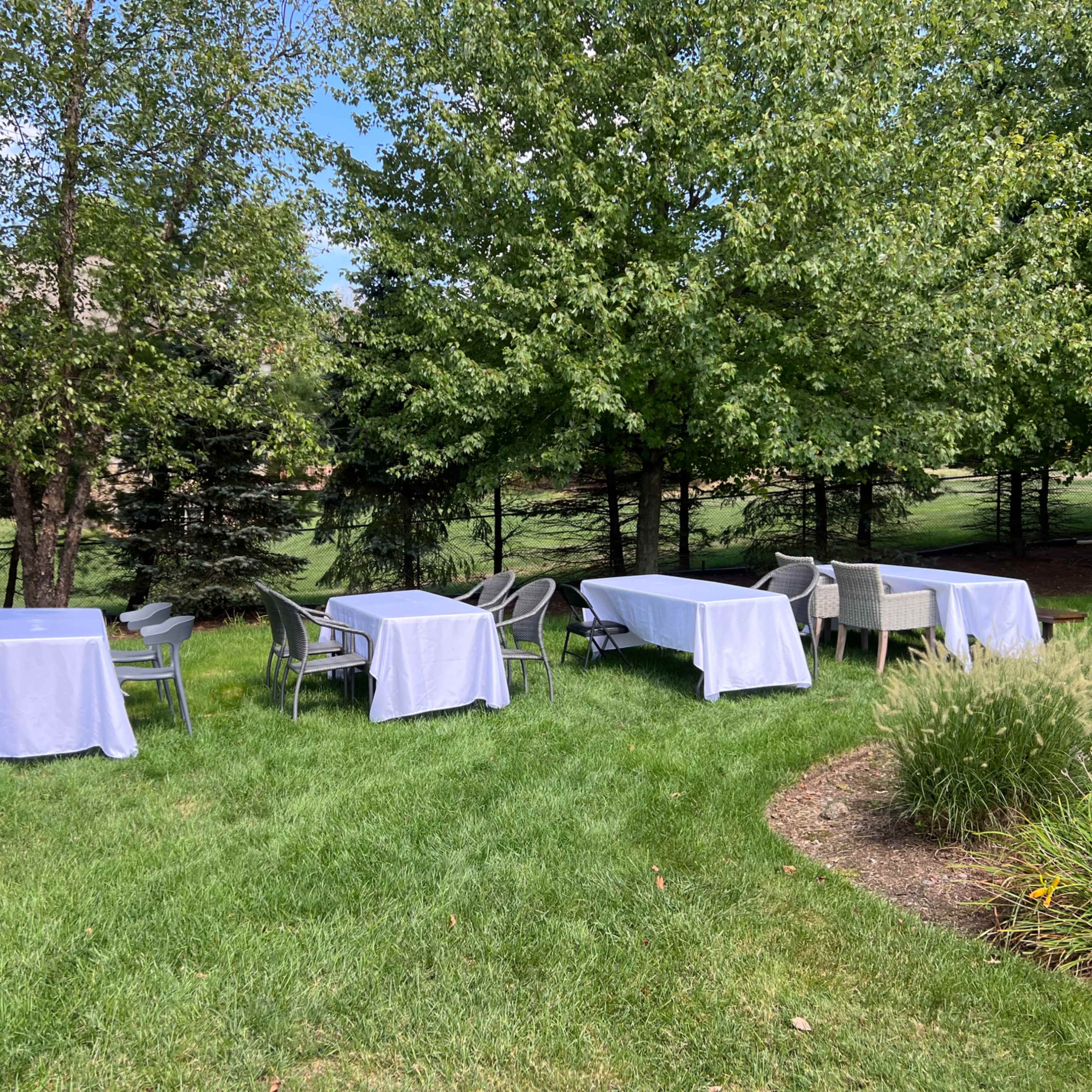 Several tables covered with white tablecloths are arranged on a grassy area surrounded by trees.