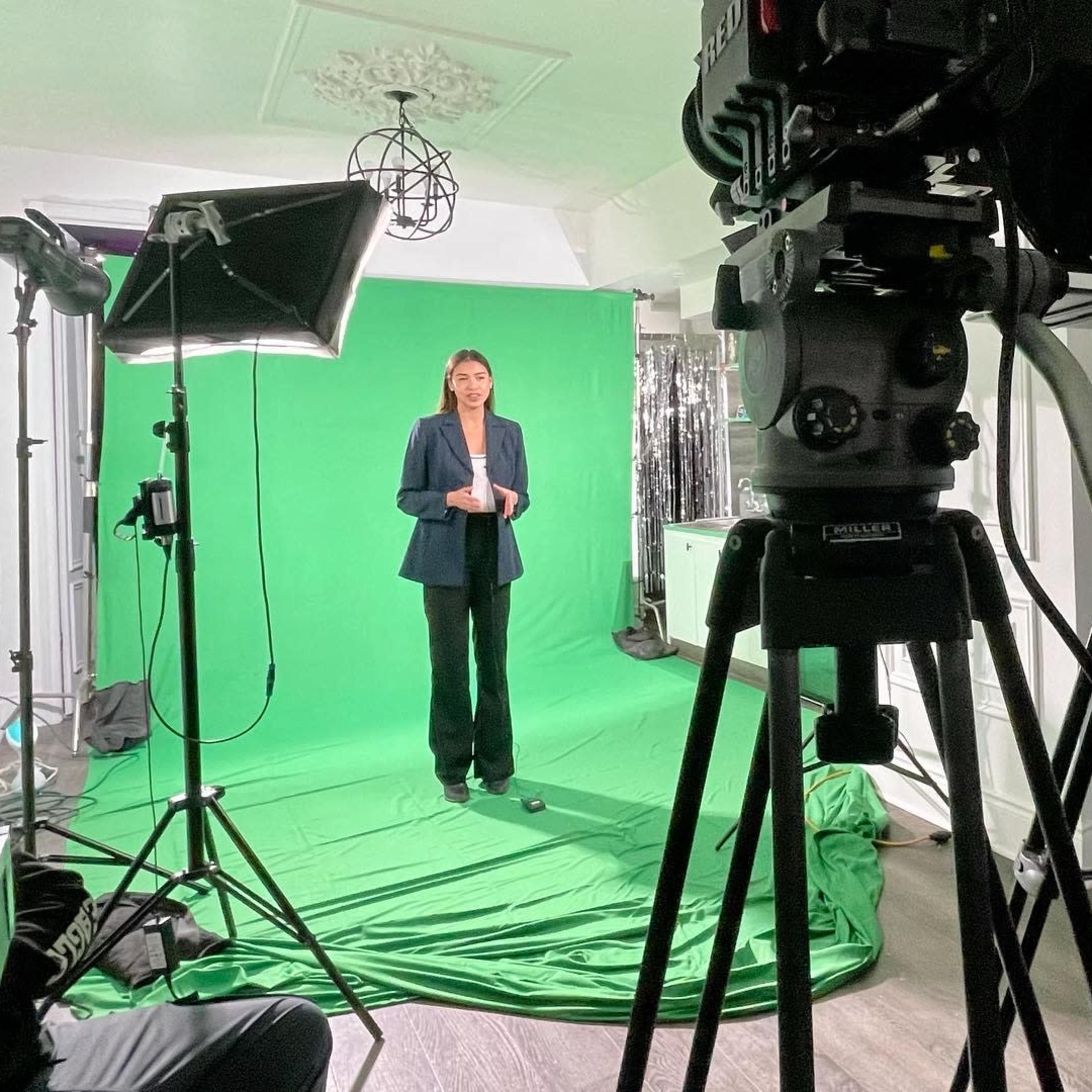 A woman stands in front of a green screen, flanked by studio lighting and cameras on tripods.