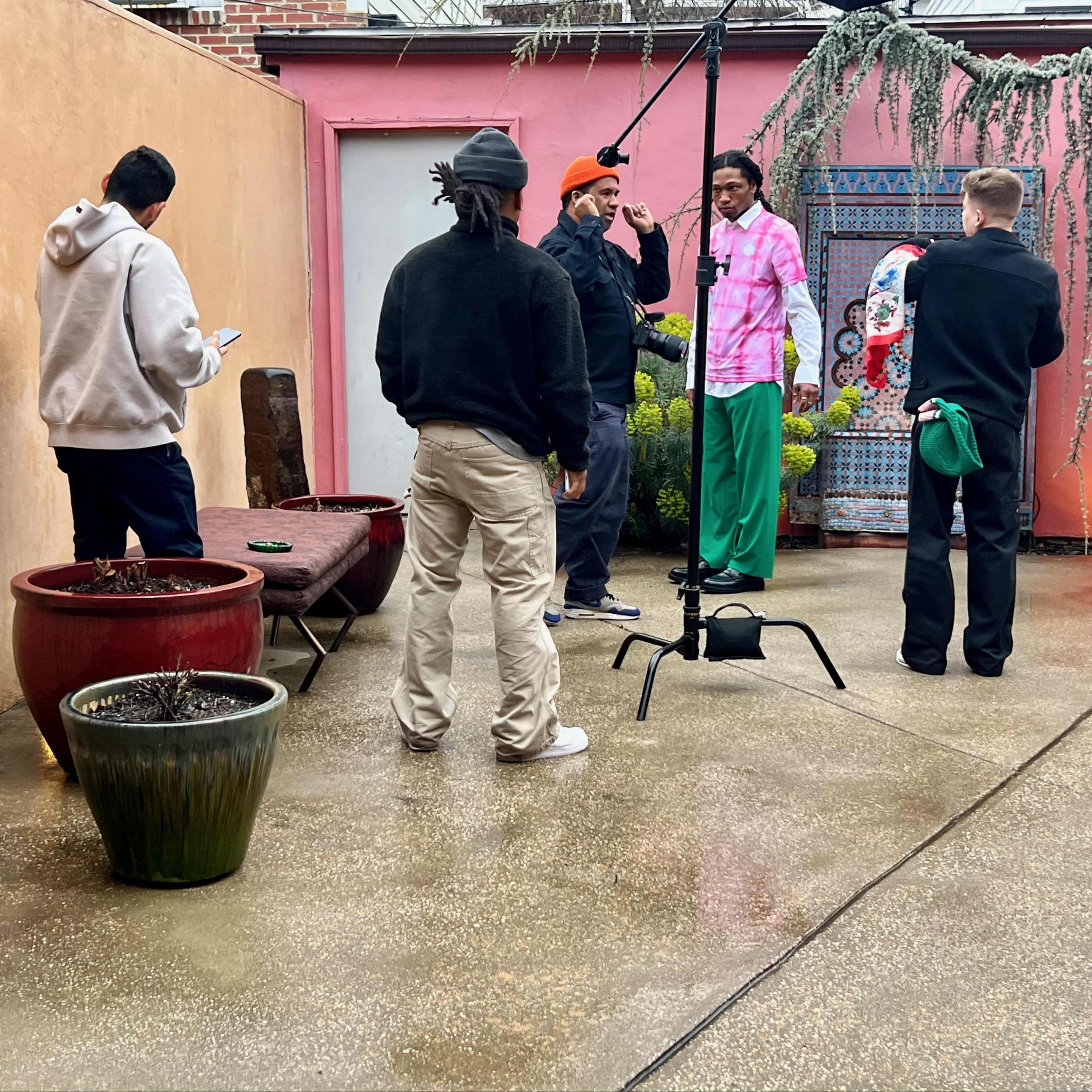 A group of five individuals stands in a courtyard with a pink wall, surrounded by potted plants, as one person poses for a photo shoot.