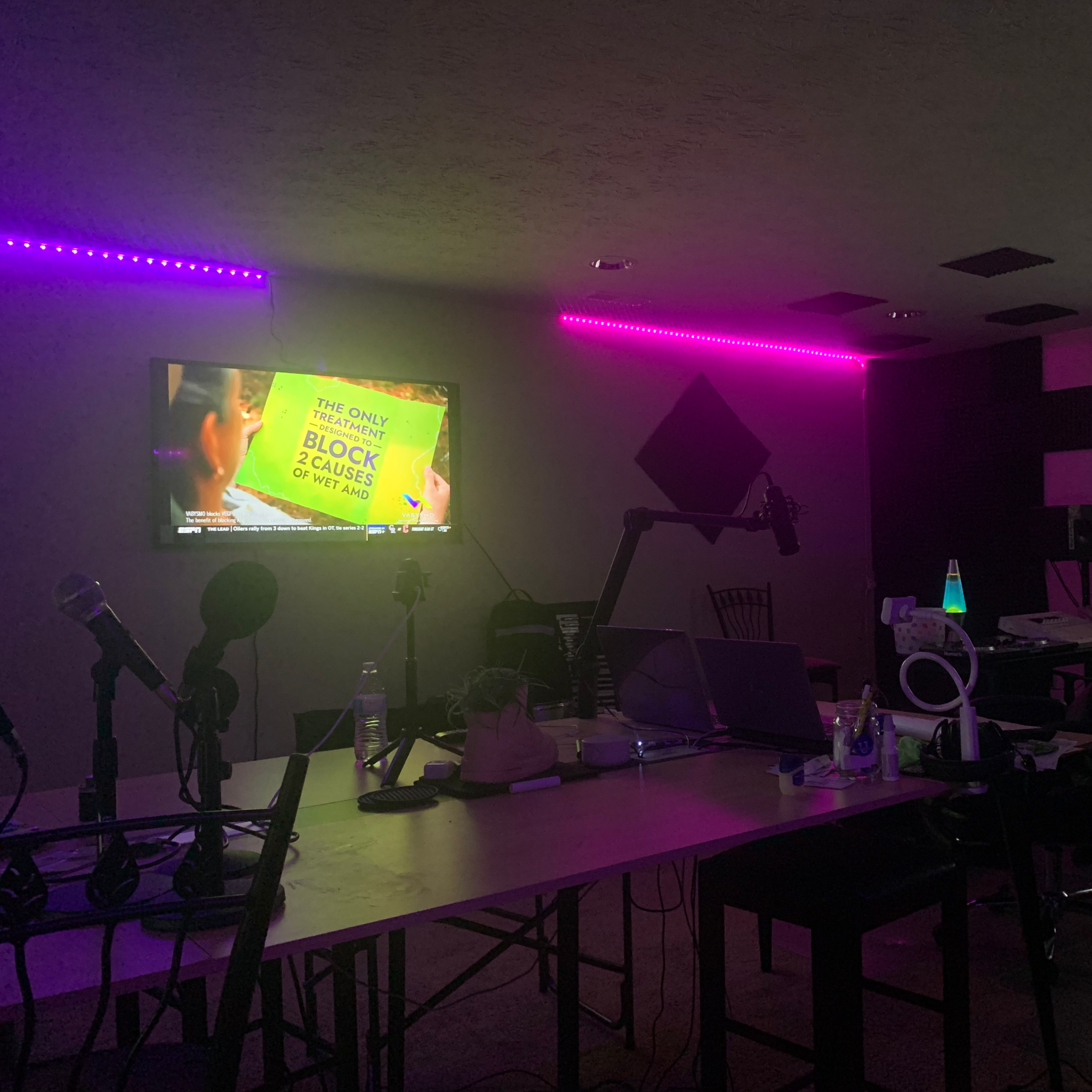 A dimly lit studio with purple neon lights, featuring a television screen, microphones, and a table with various equipment.