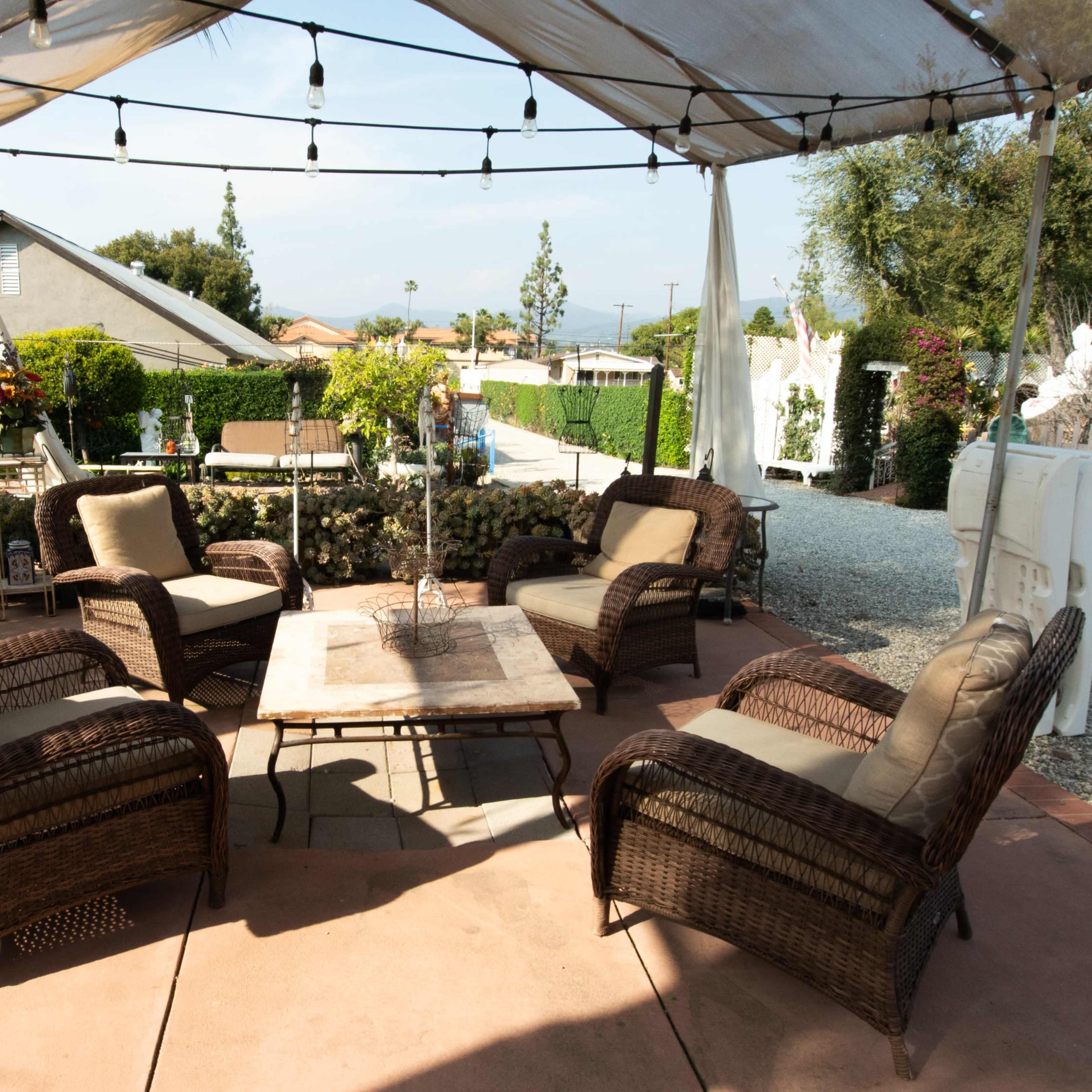 A covered outdoor seating area features four wicker chairs arranged around a central table, surrounded by greenery and decorative elements.