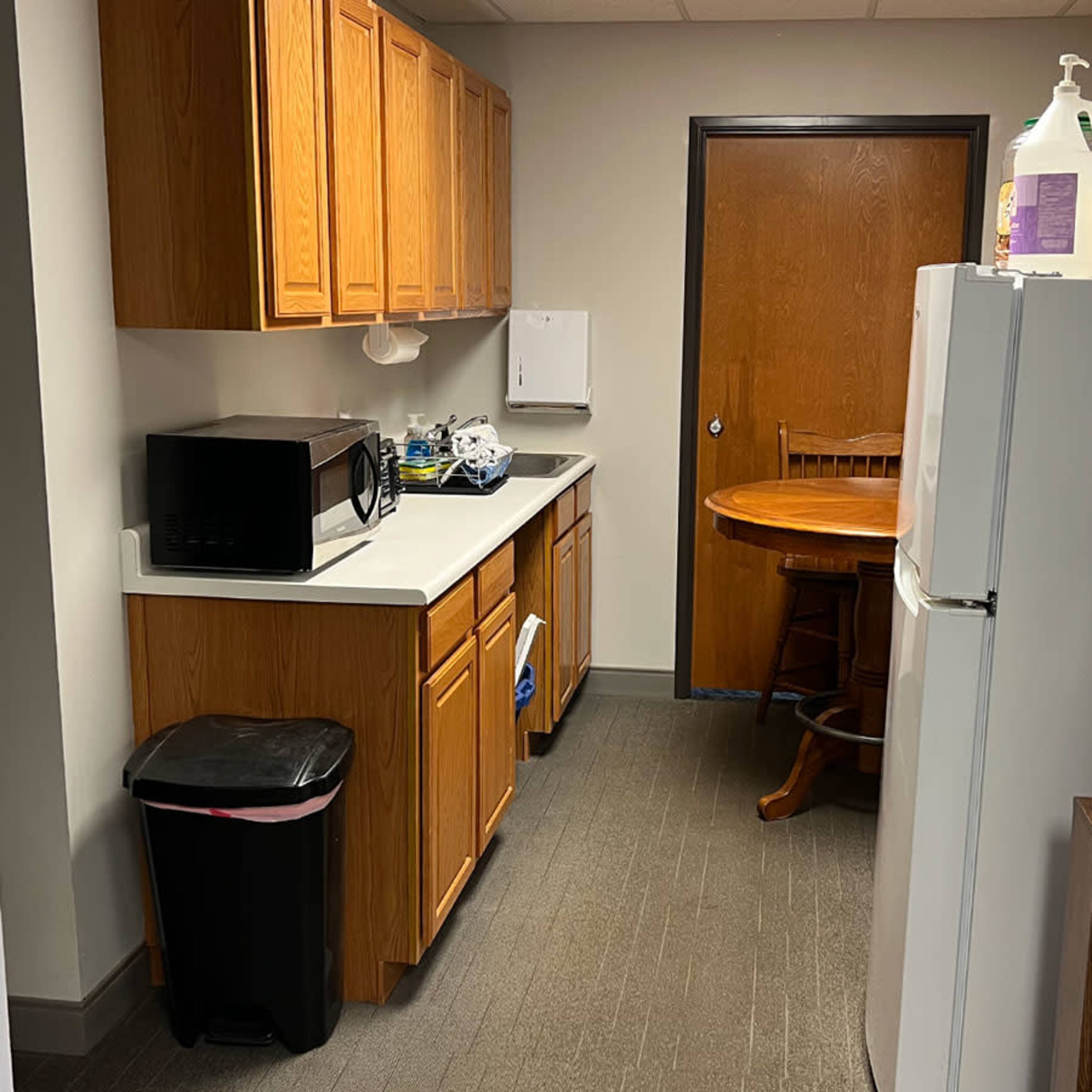 A small kitchen area with wooden cabinets, a microwave, a round wooden table, and a refrigerator beside a water cooler.