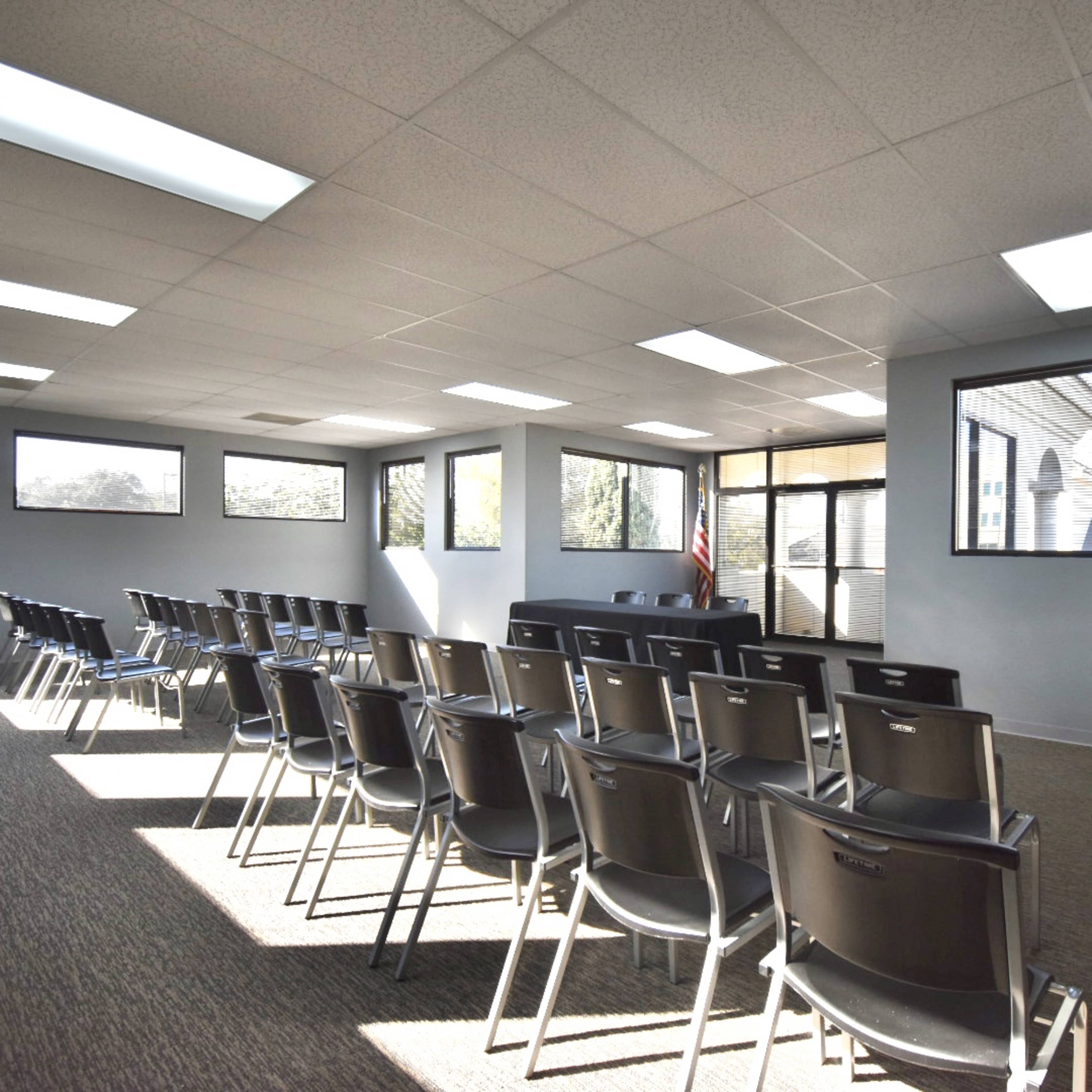 A vacant meeting room features rows of black chairs facing a front area with large windows and a door leading outside.