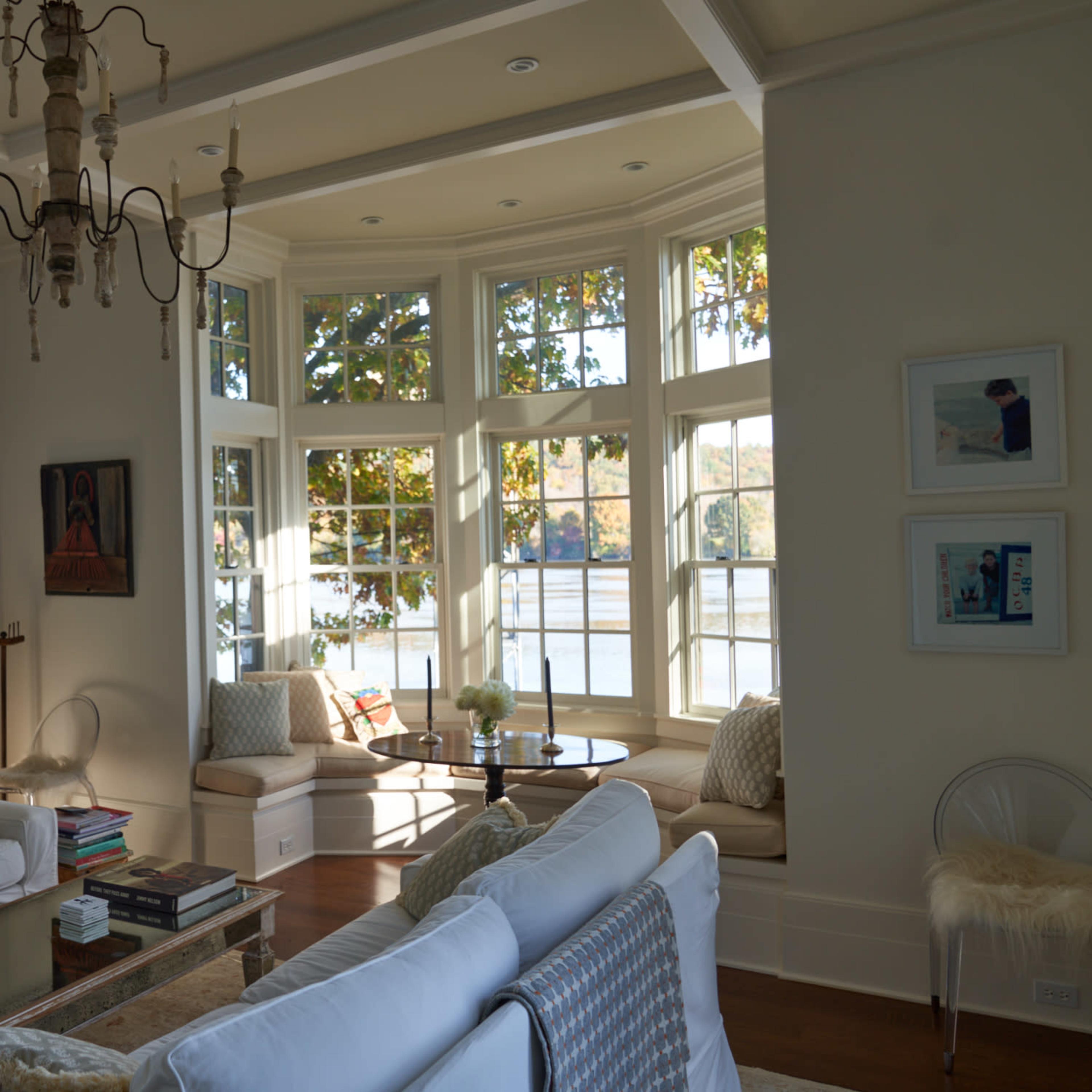 A bright living room features large windows overlooking water, with a chandelier and white sofas arranged around a coffee table.