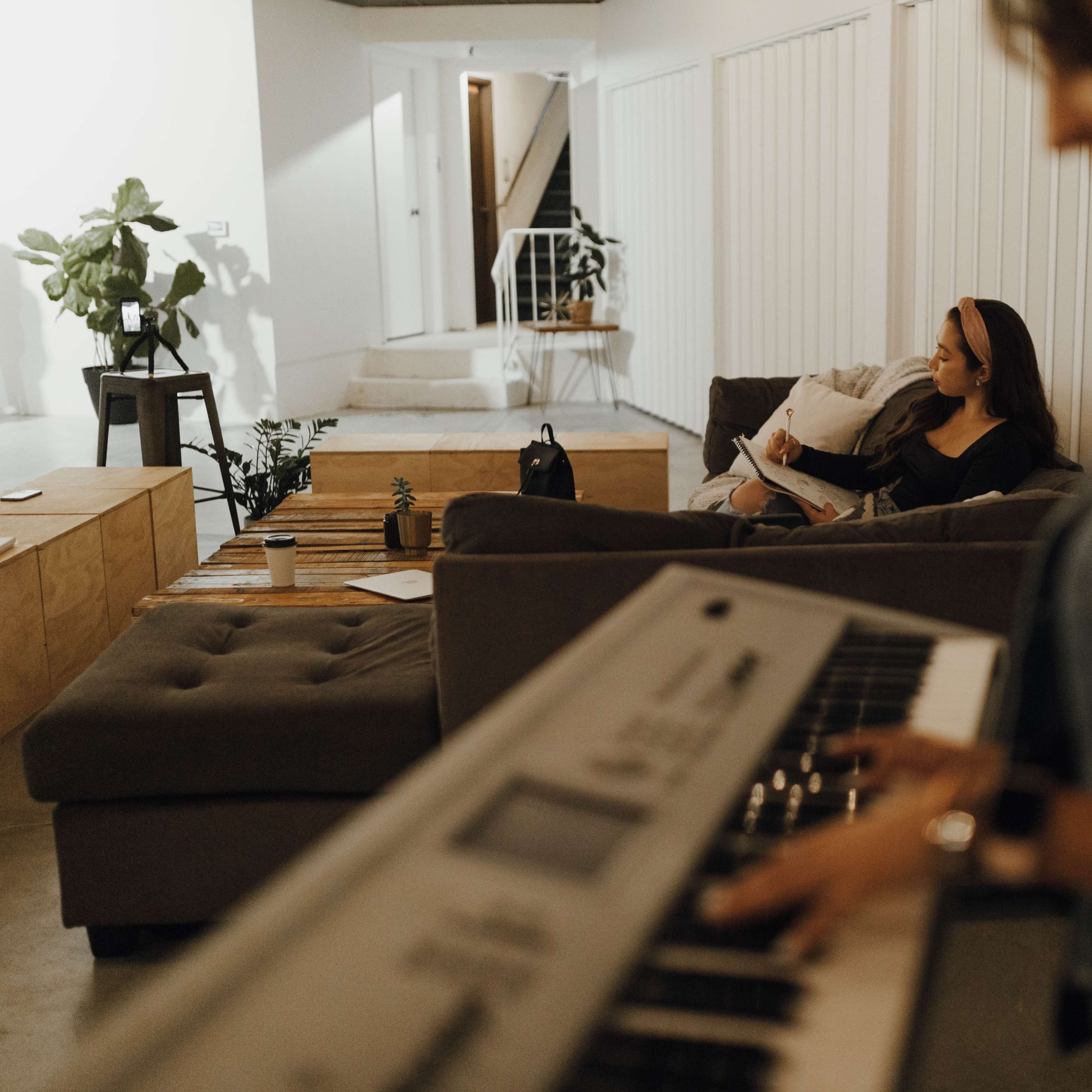 A person plays a keyboard in the foreground while another sits on a couch in the background, writing notes in a notebook.