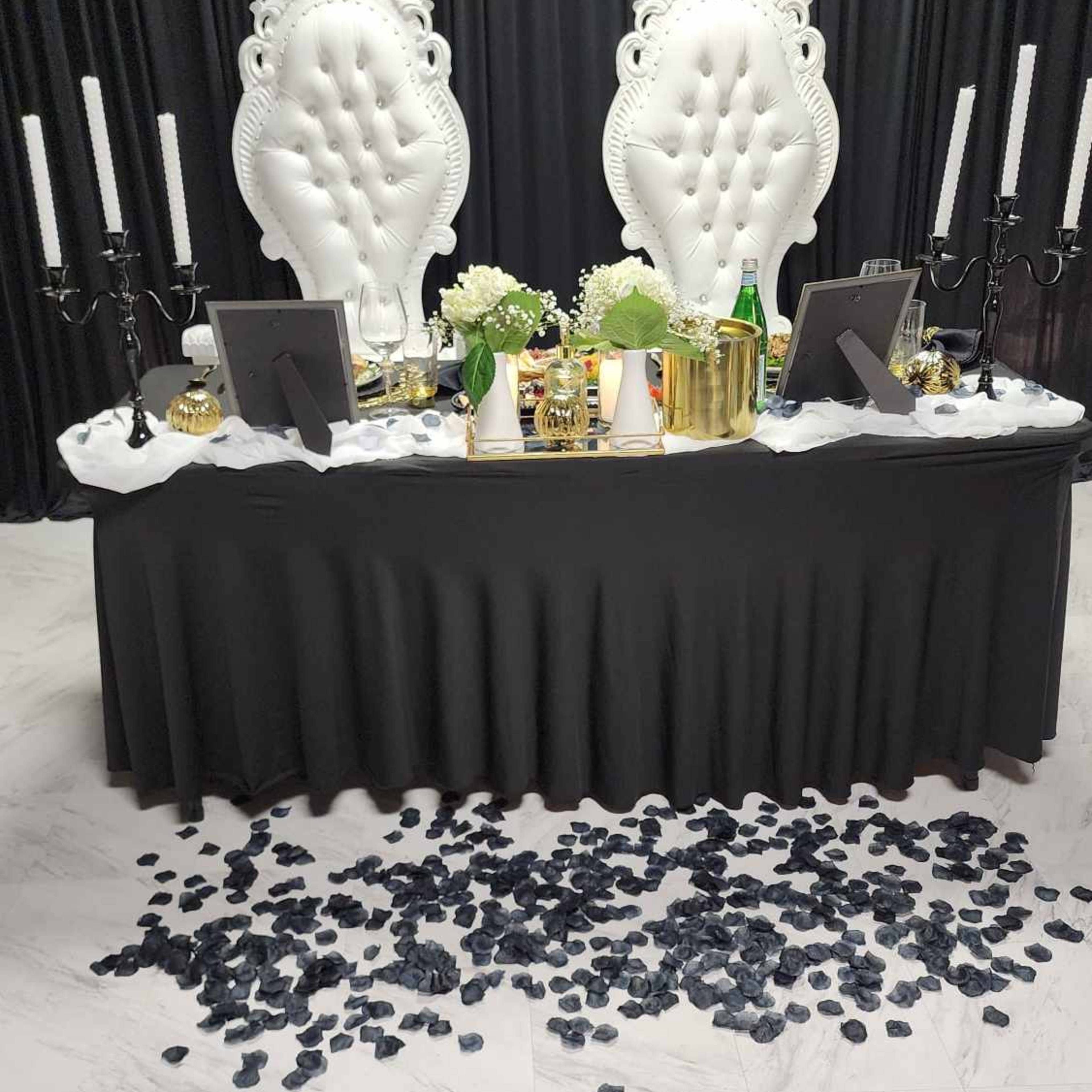 A black-topped table is set with decor and two ornate white chairs against a black backdrop, with black rose petals scattered on the floor.