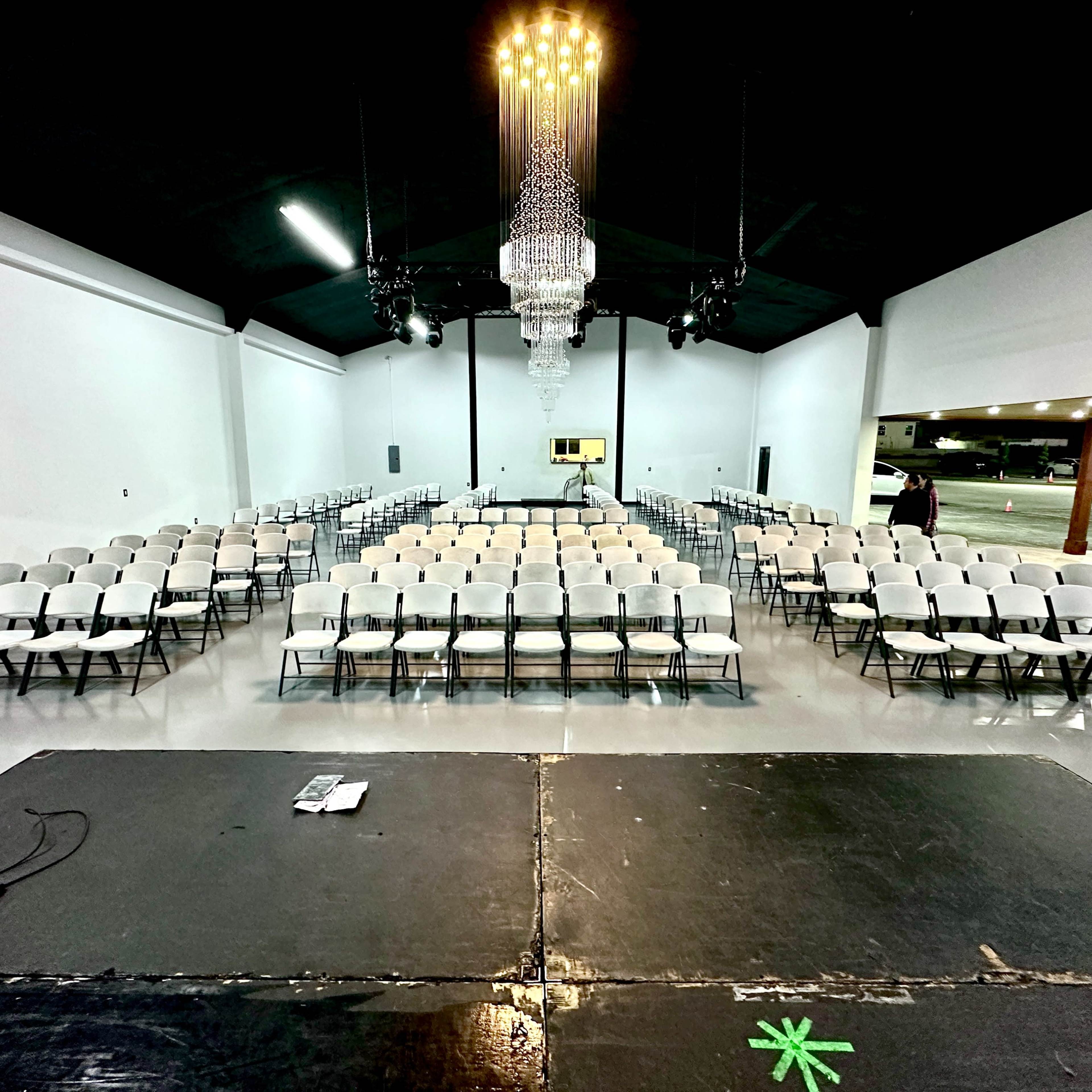 A spacious indoor venue set up with rows of white folding chairs facing a stage, illuminated by a chandelier overhead.