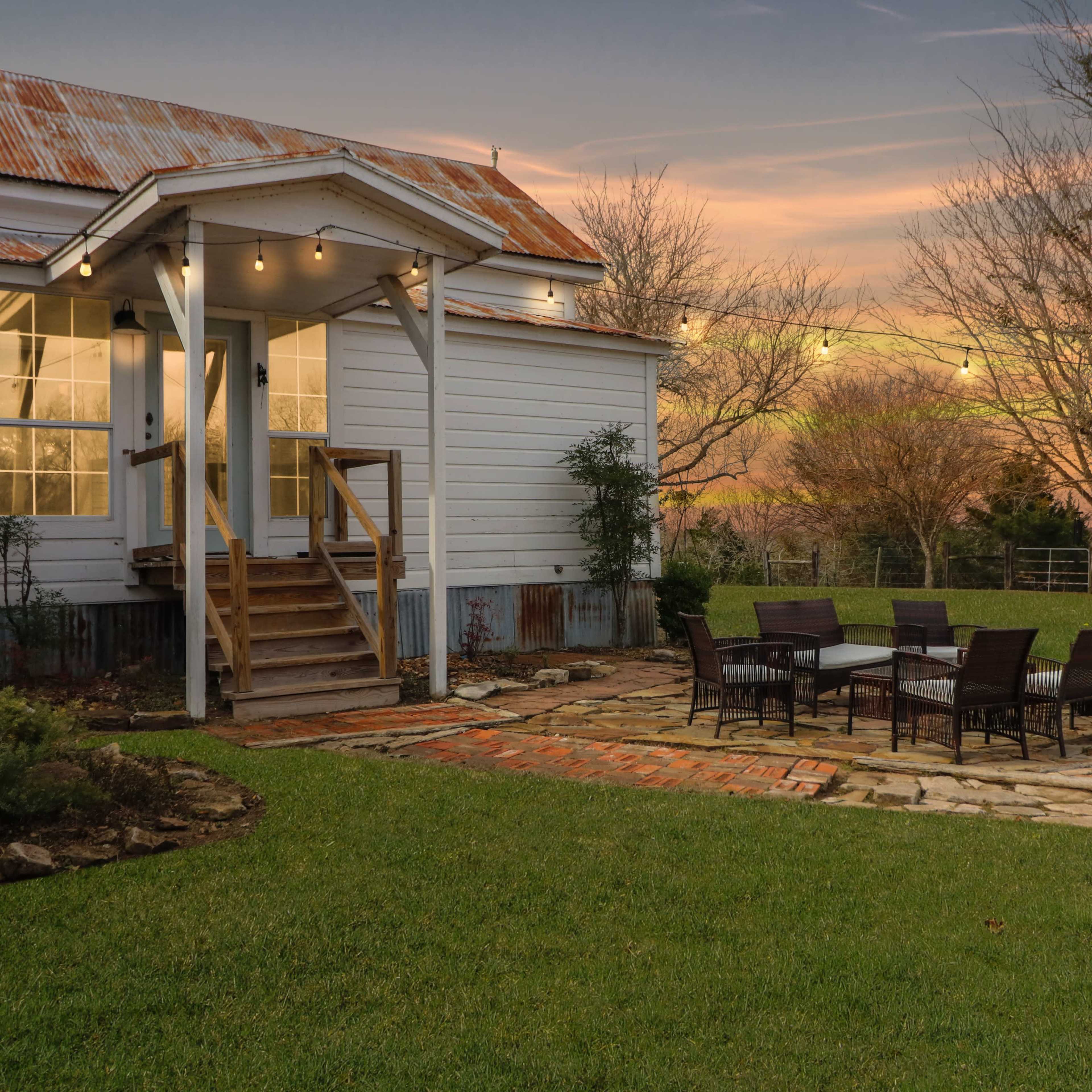 A house with a covered porch, a brick pathway, and a patio area with a stone seating arrangement in a grassy yard at dusk.