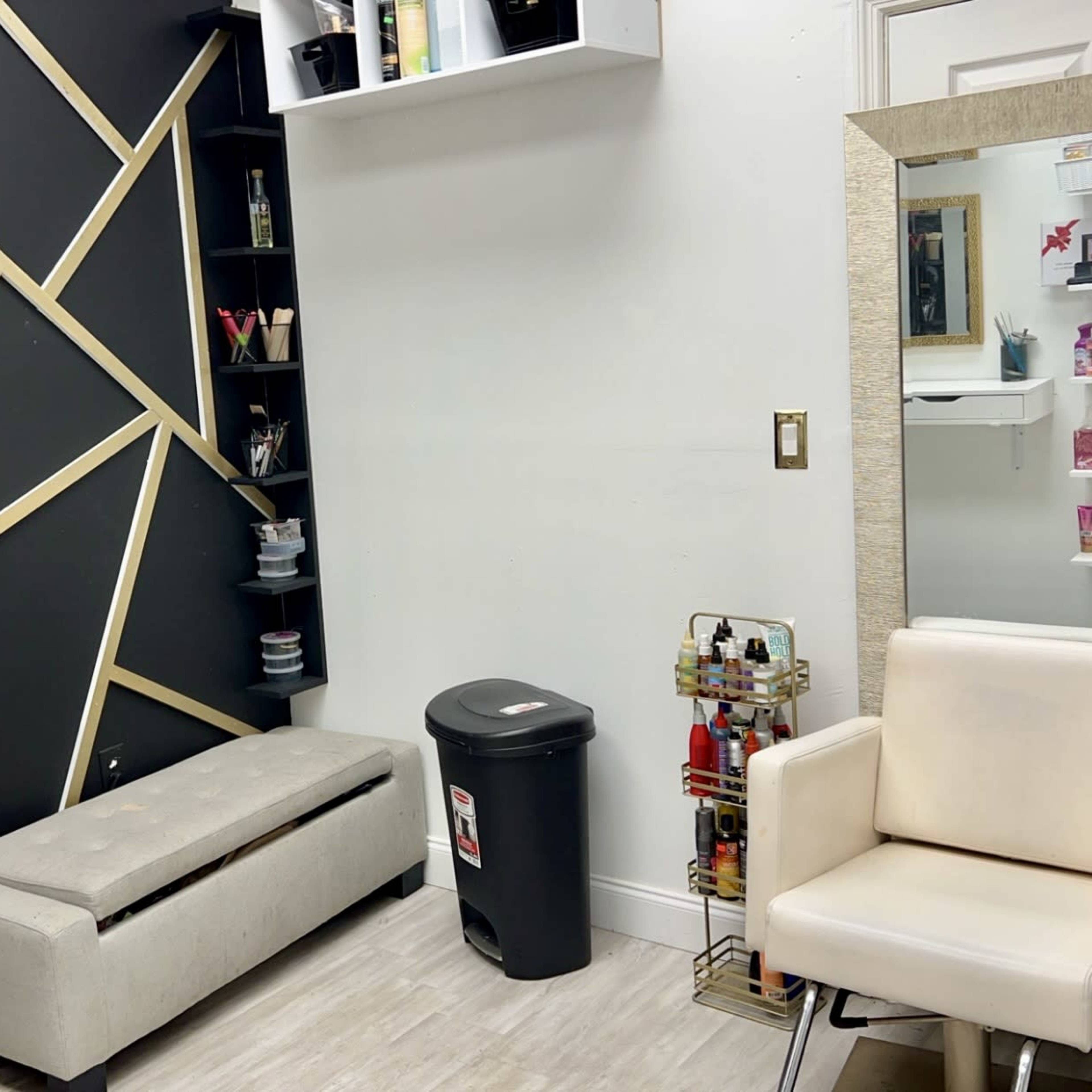 A minimalist salon space featuring a black and gold decorative wall, a white mirror, a gray bench, a white chair, and shelving with cosmetic products.