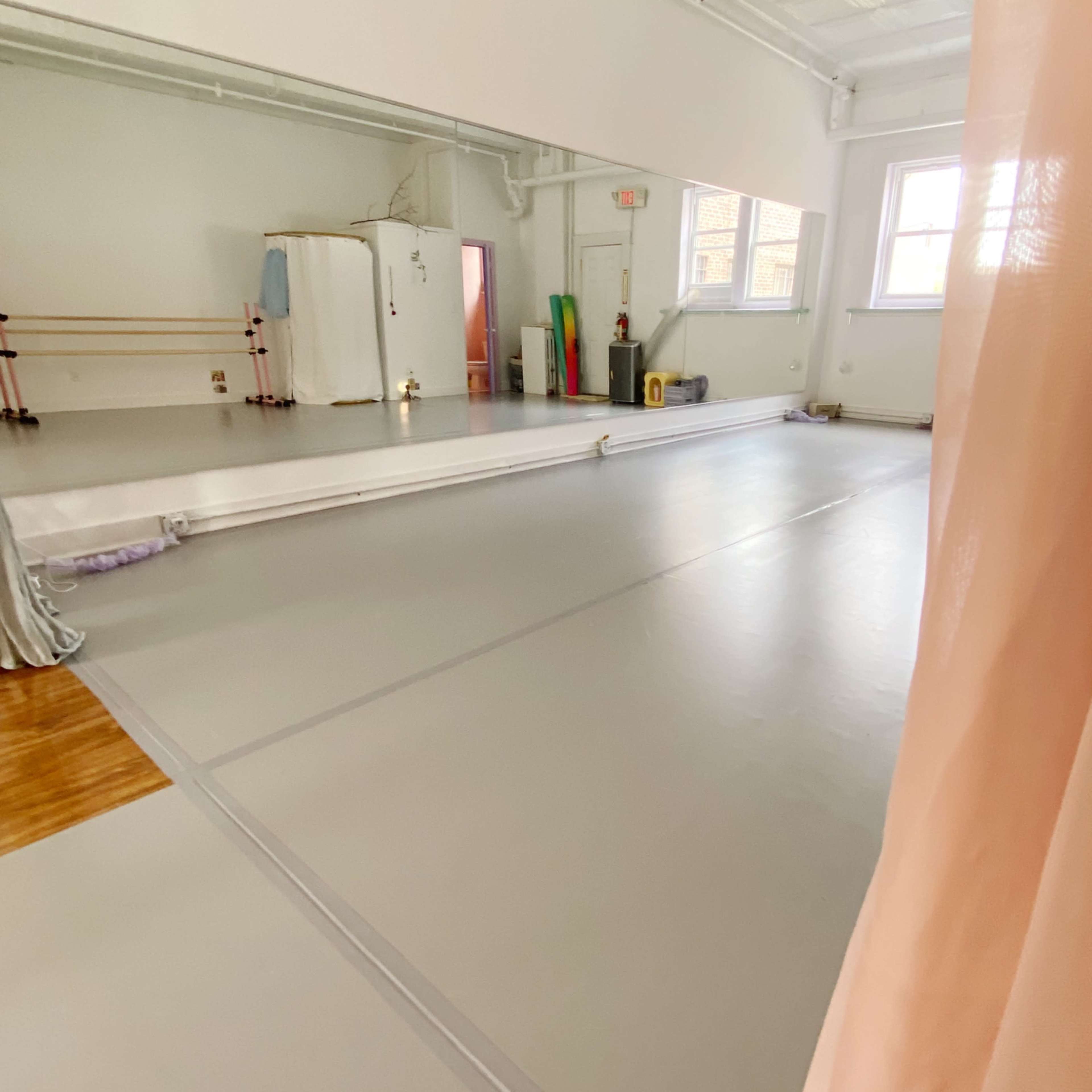 A spacious dance studio with a mirrored wall, wooden ballet barres, and a gray floor.
