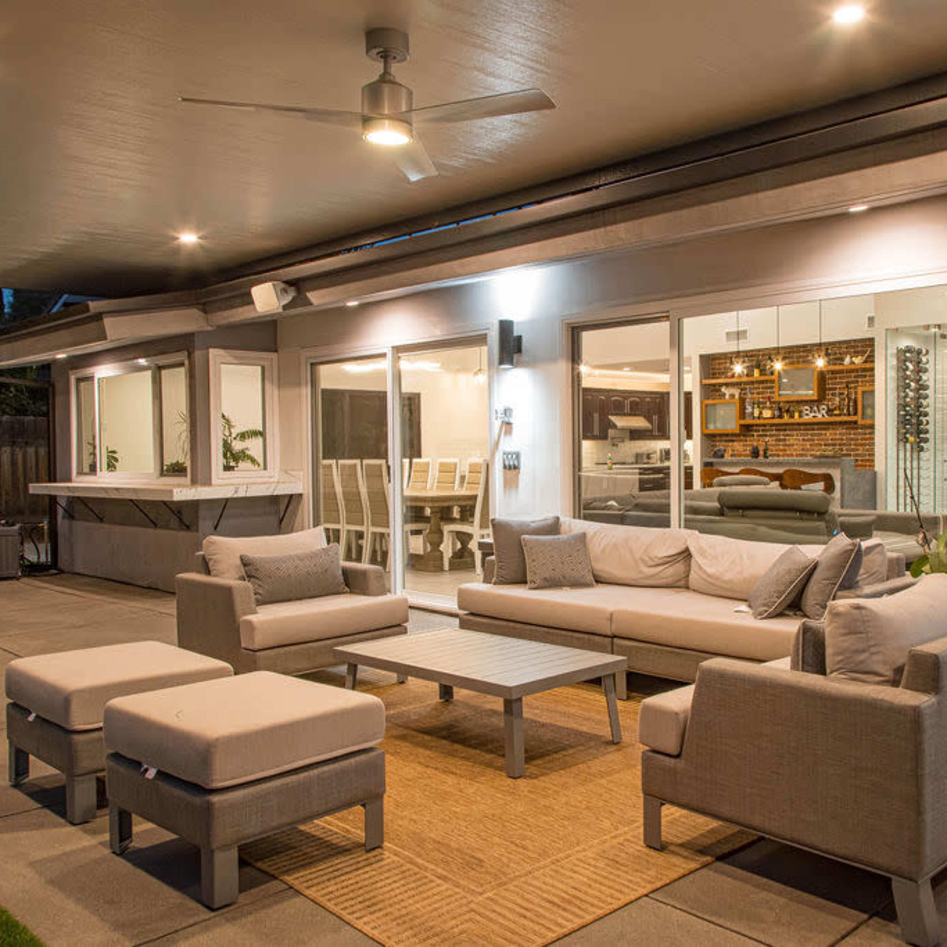 A modern outdoor seating area features comfortable gray furniture arranged around a coffee table, overlooking large windows that provide a view of an interior dining space.