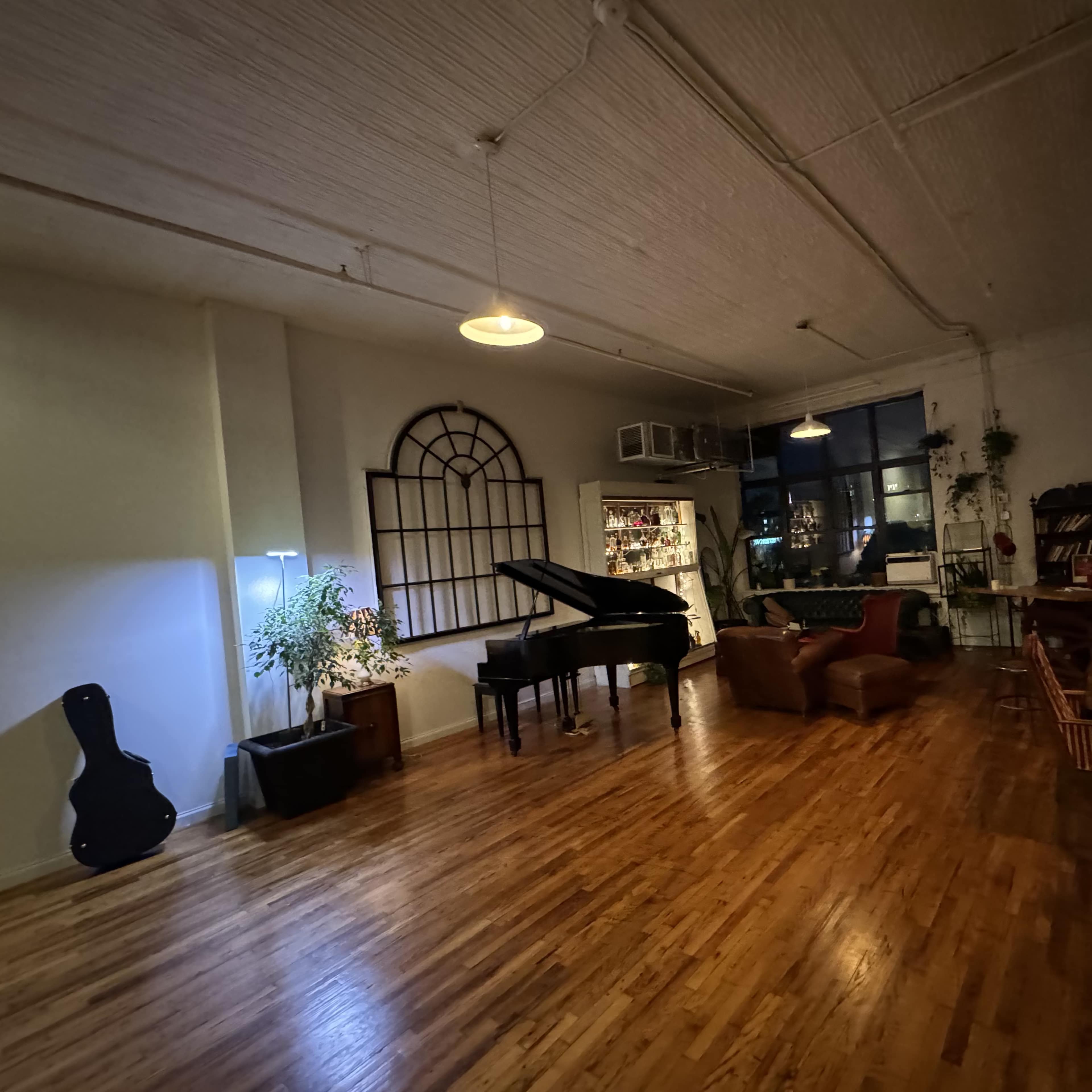 A spacious, dimly lit room featuring a grand piano, a guitar resting against a plant, and various pieces of furniture along the walls.