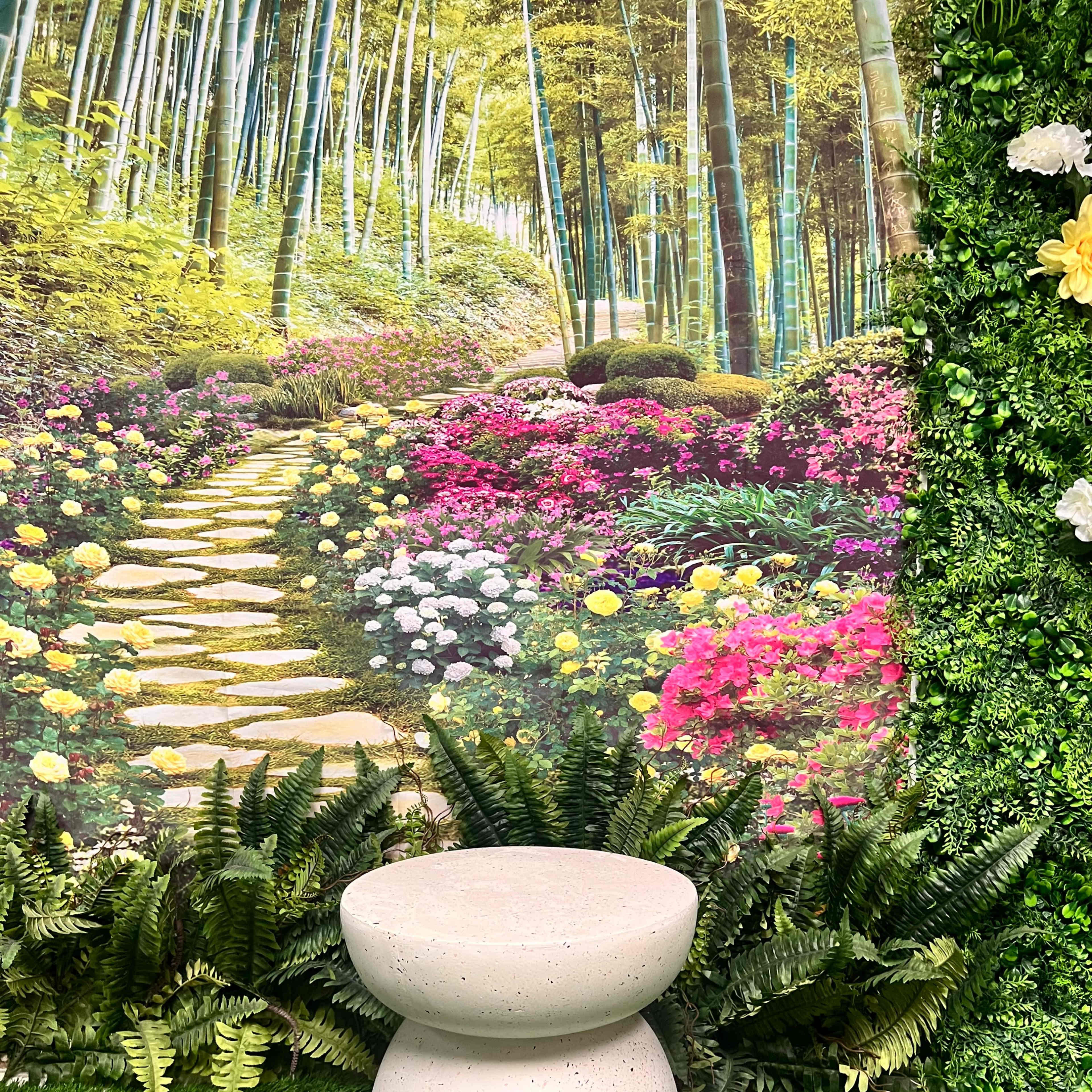 A round, textured concrete stool stands in front of a vibrant mural depicting a garden path lined with flowers and bamboo.