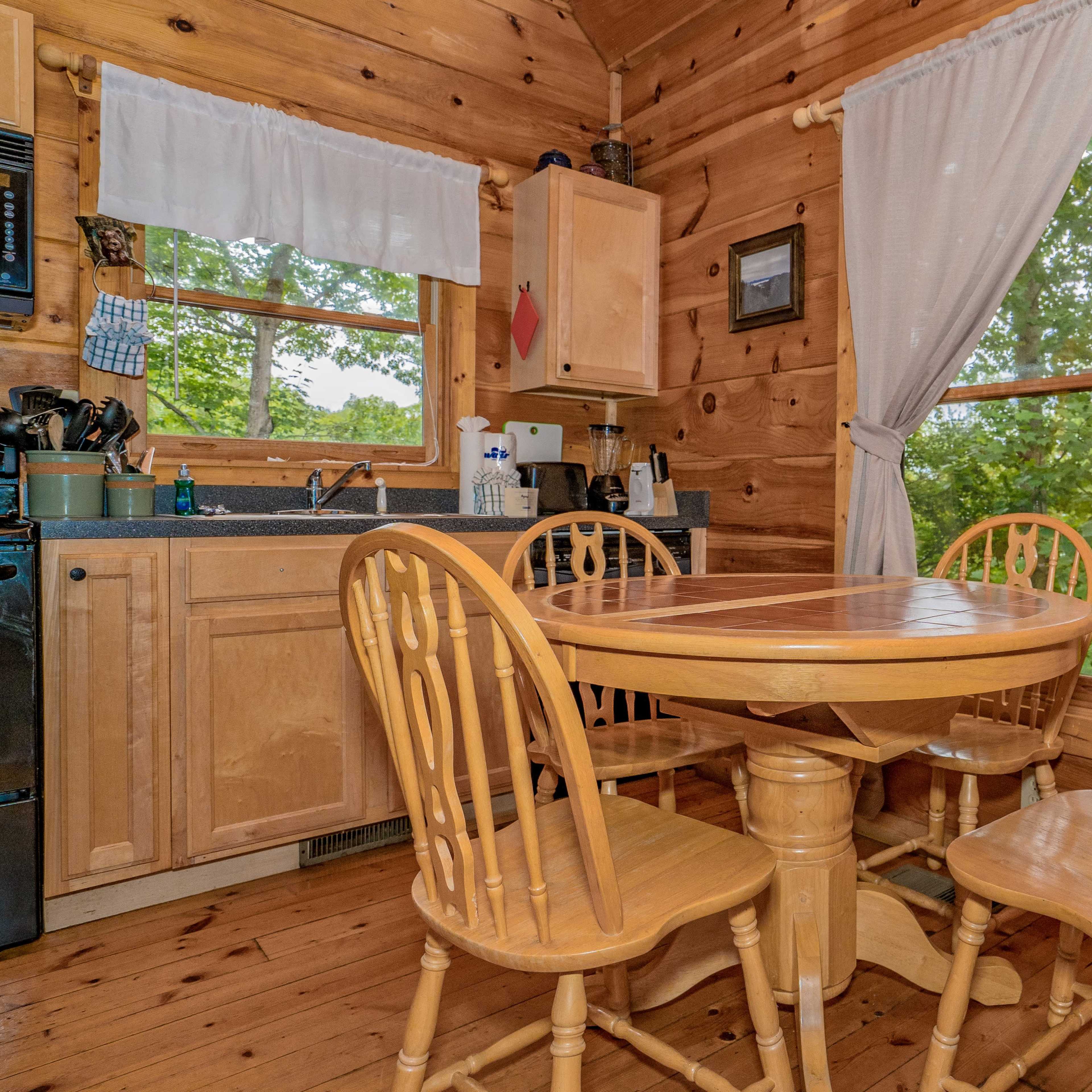 A wooden kitchen features a round table surrounded by four chairs, with a sink and cabinets visible in the background.