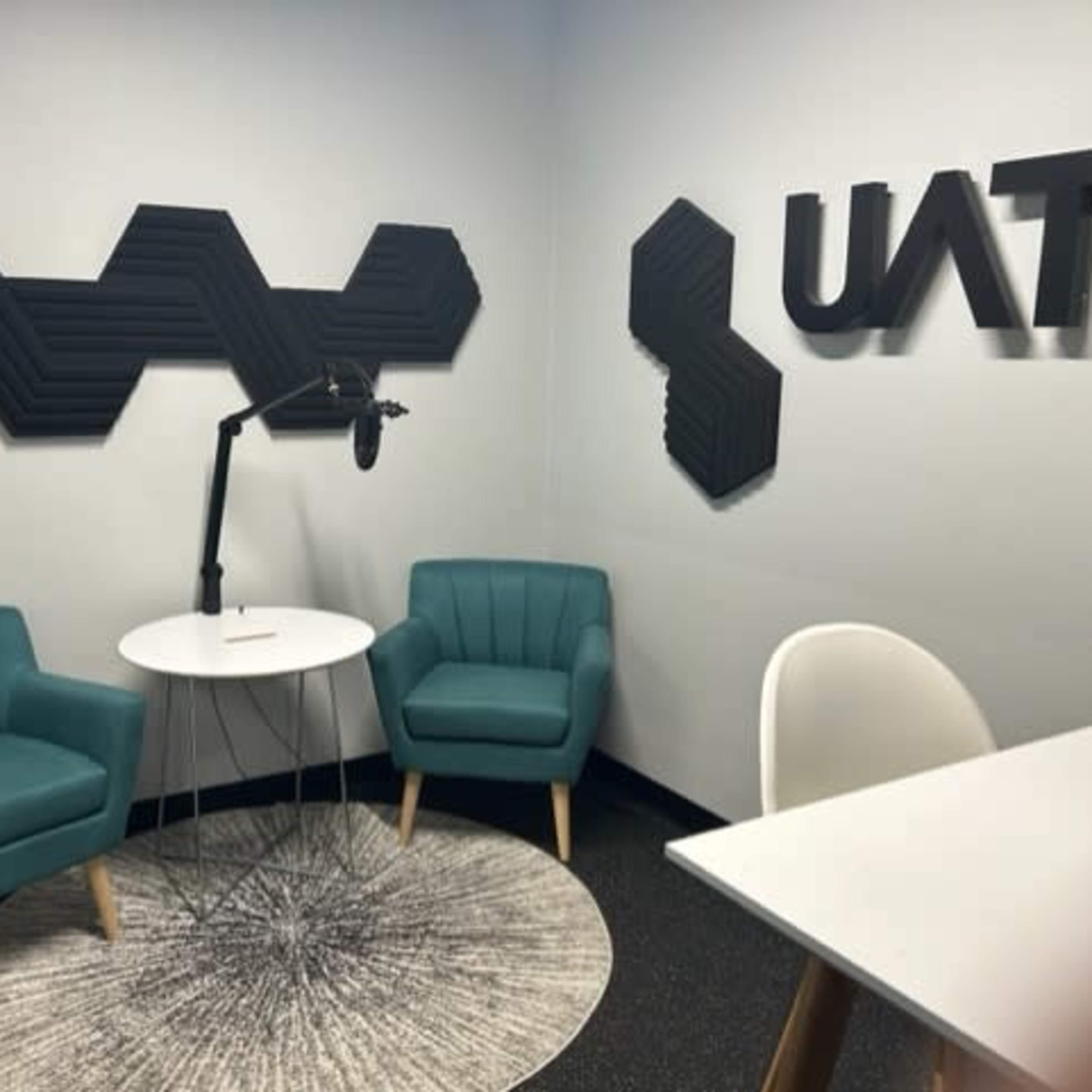 A modern room with two teal chairs, a round table, and a microphone stand, featuring acoustic wall panels and the letters "UAT" on the wall.