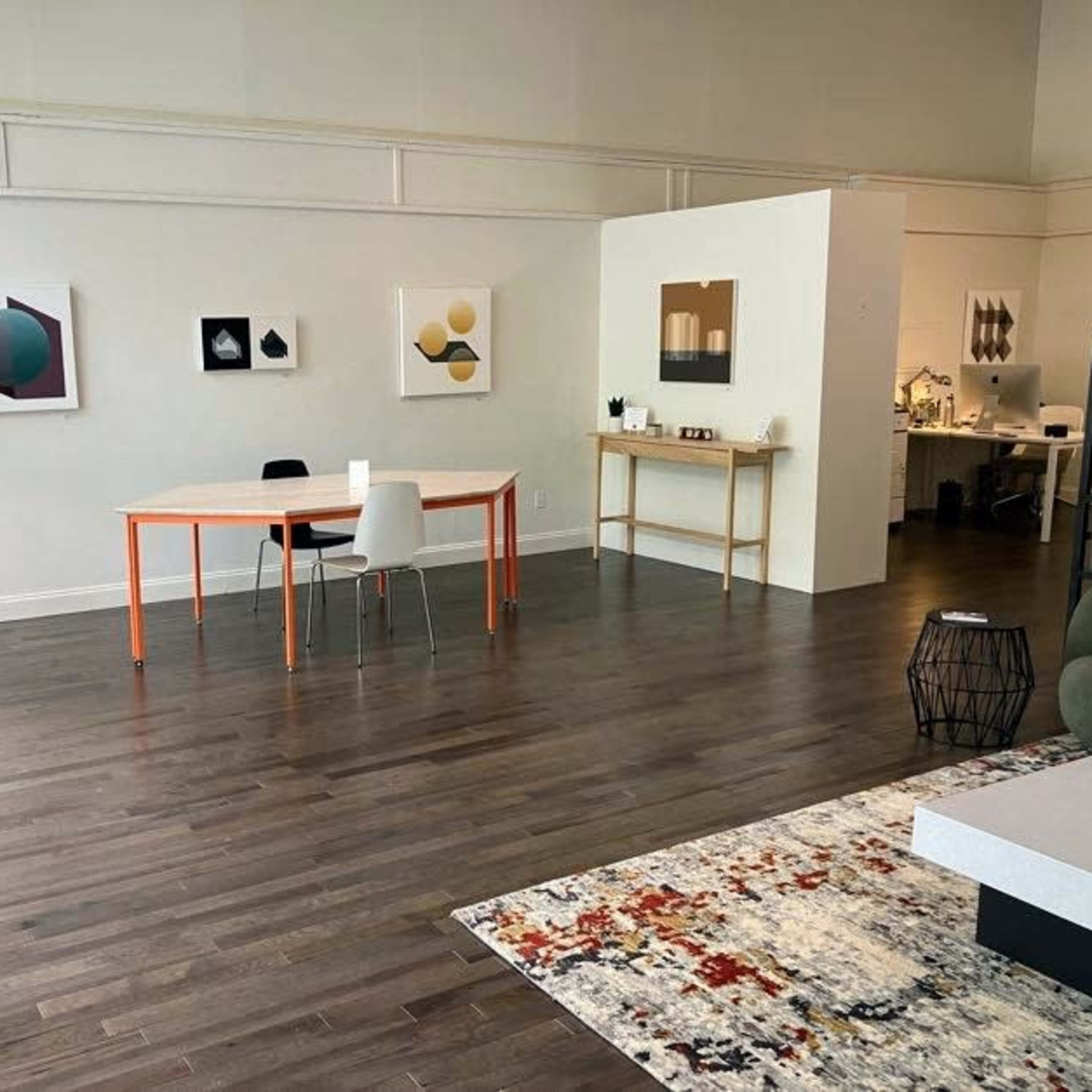 An art gallery space with wooden flooring, featuring abstract artworks on the walls and a couple of tables with chairs arranged in the foreground.