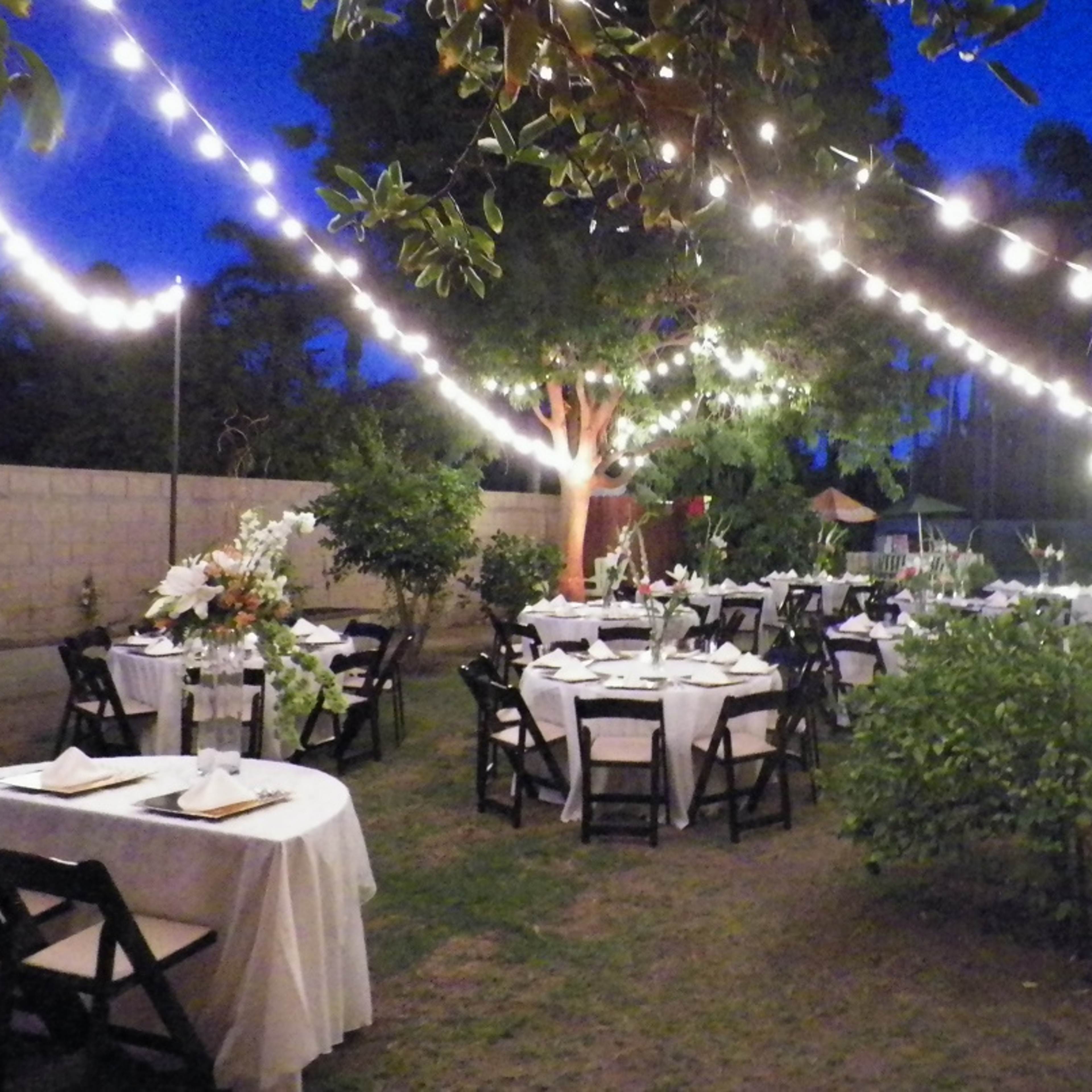 The scene depicts an outdoor dining area with several tables set for a meal, illuminated by string lights amid trees and greenery.