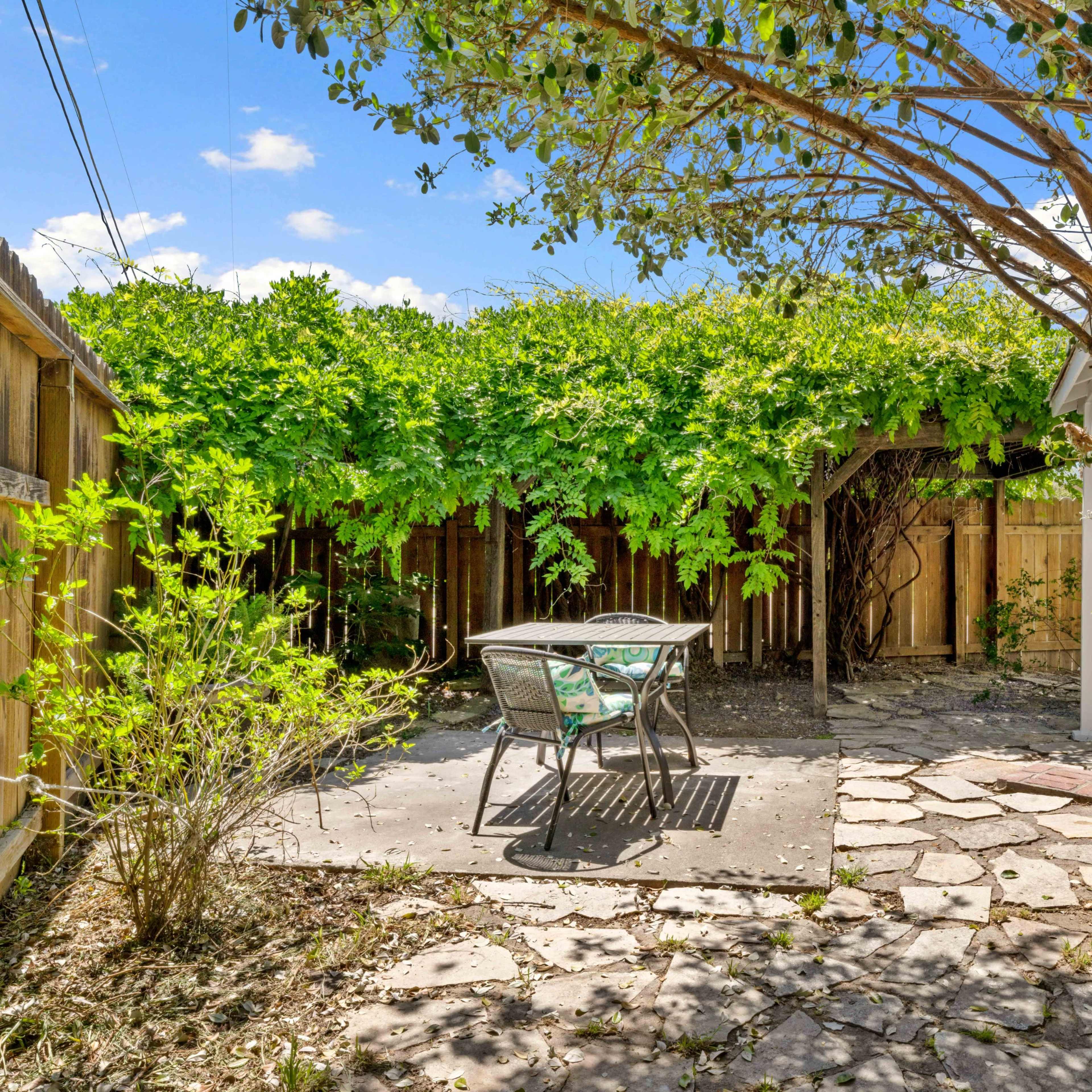 A small stone patio with a metal table and chairs is surrounded by dense greenery and wooden fencing.