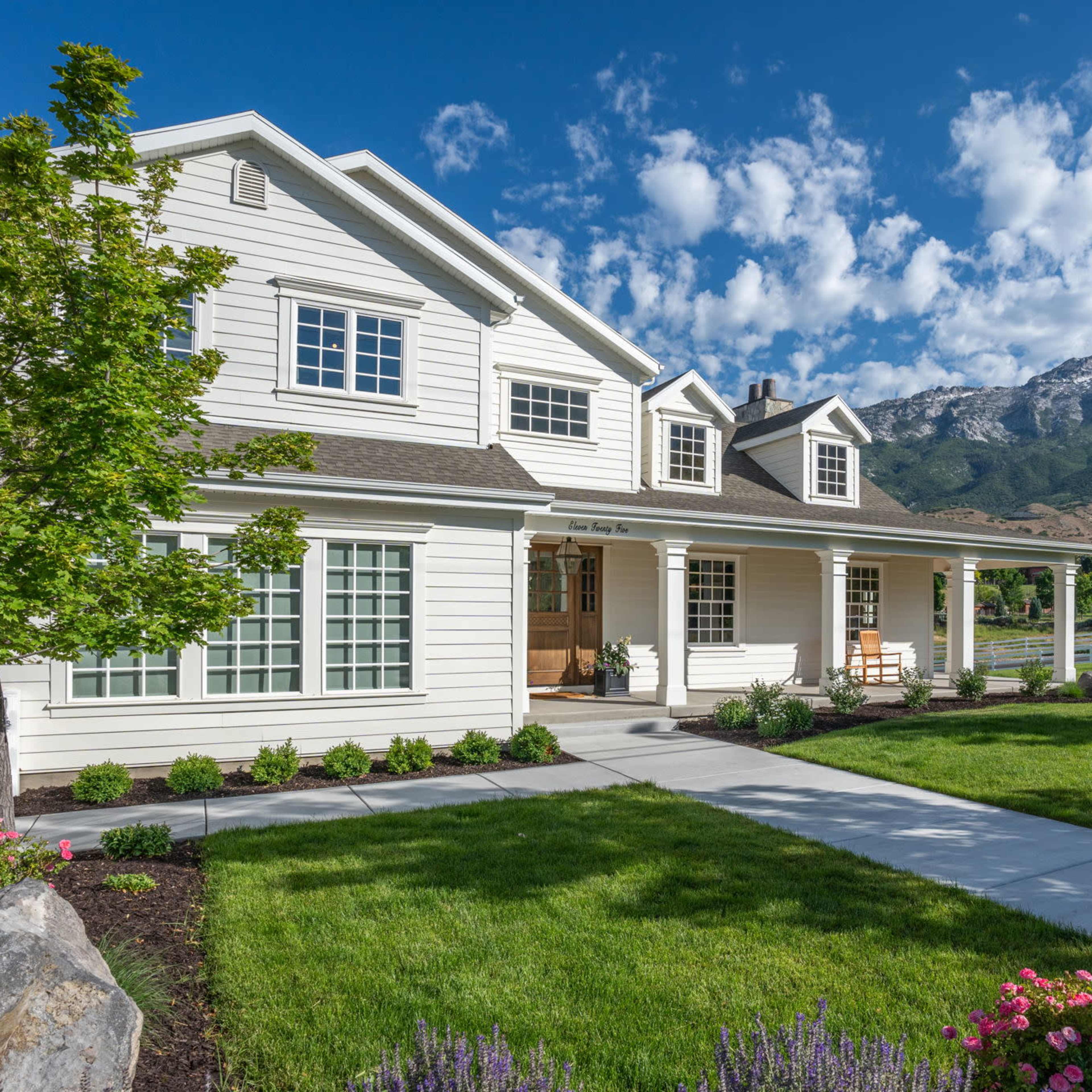 A well-maintained two-story white house with a large front porch is set against a backdrop of mountains and a blue sky.