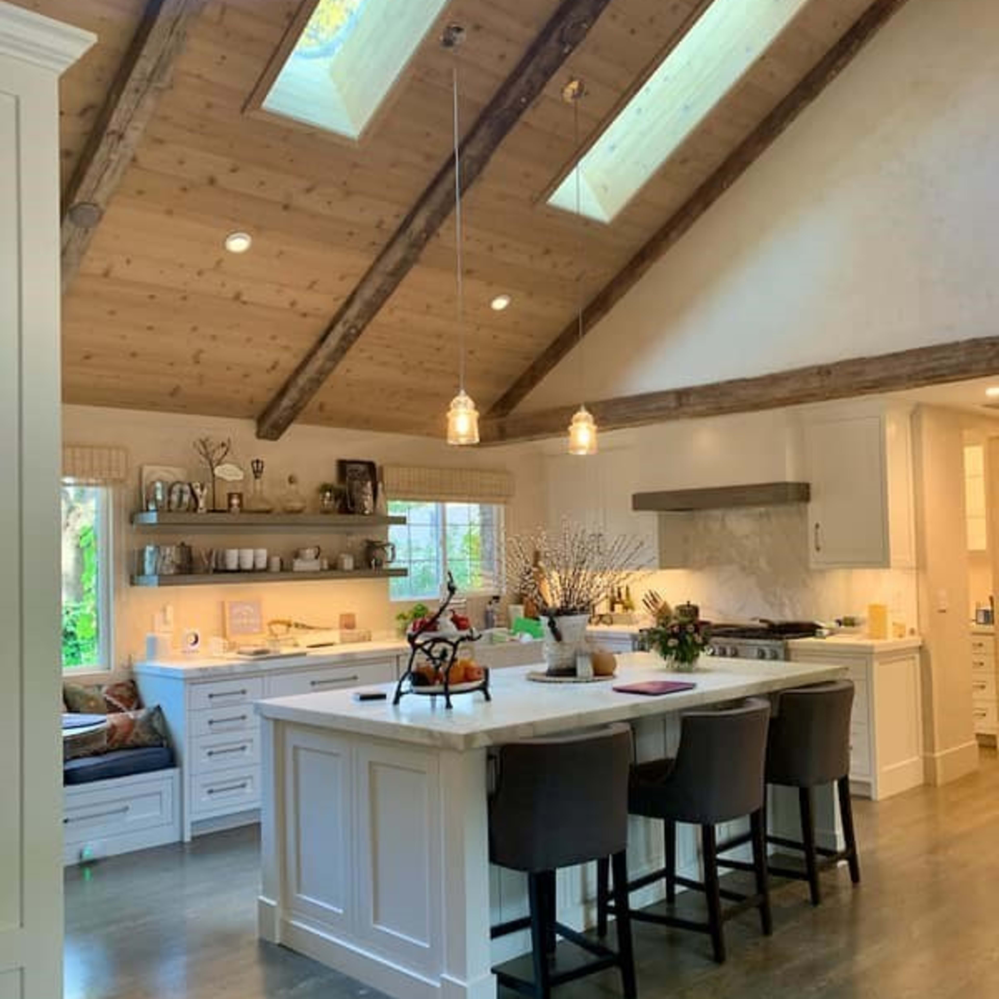 A modern kitchen with a central island, wooden beams, skylights, and a cozy seating area by a window.