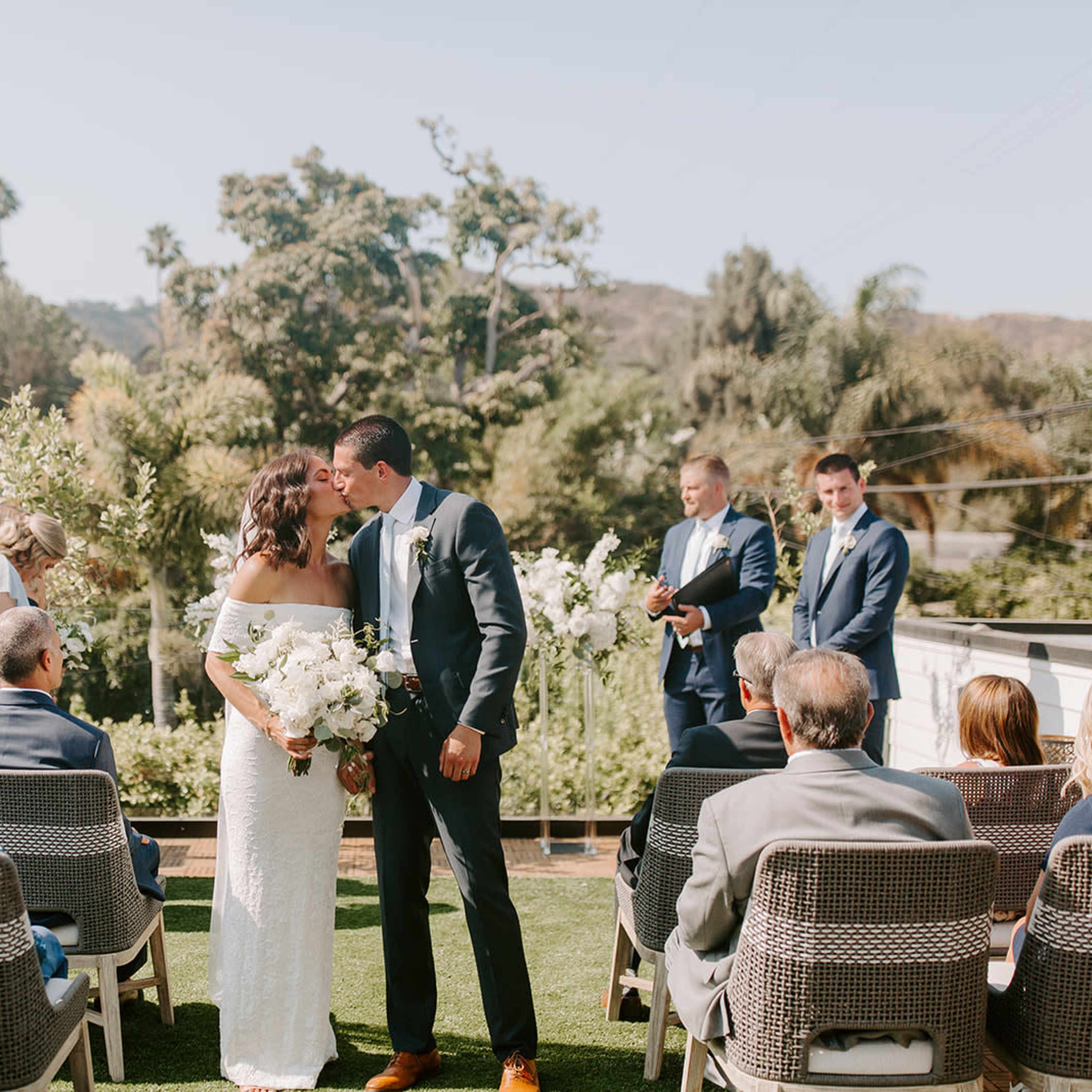 A couple shares a kiss during their outdoor wedding ceremony, surrounded by guests seated on a lawn.