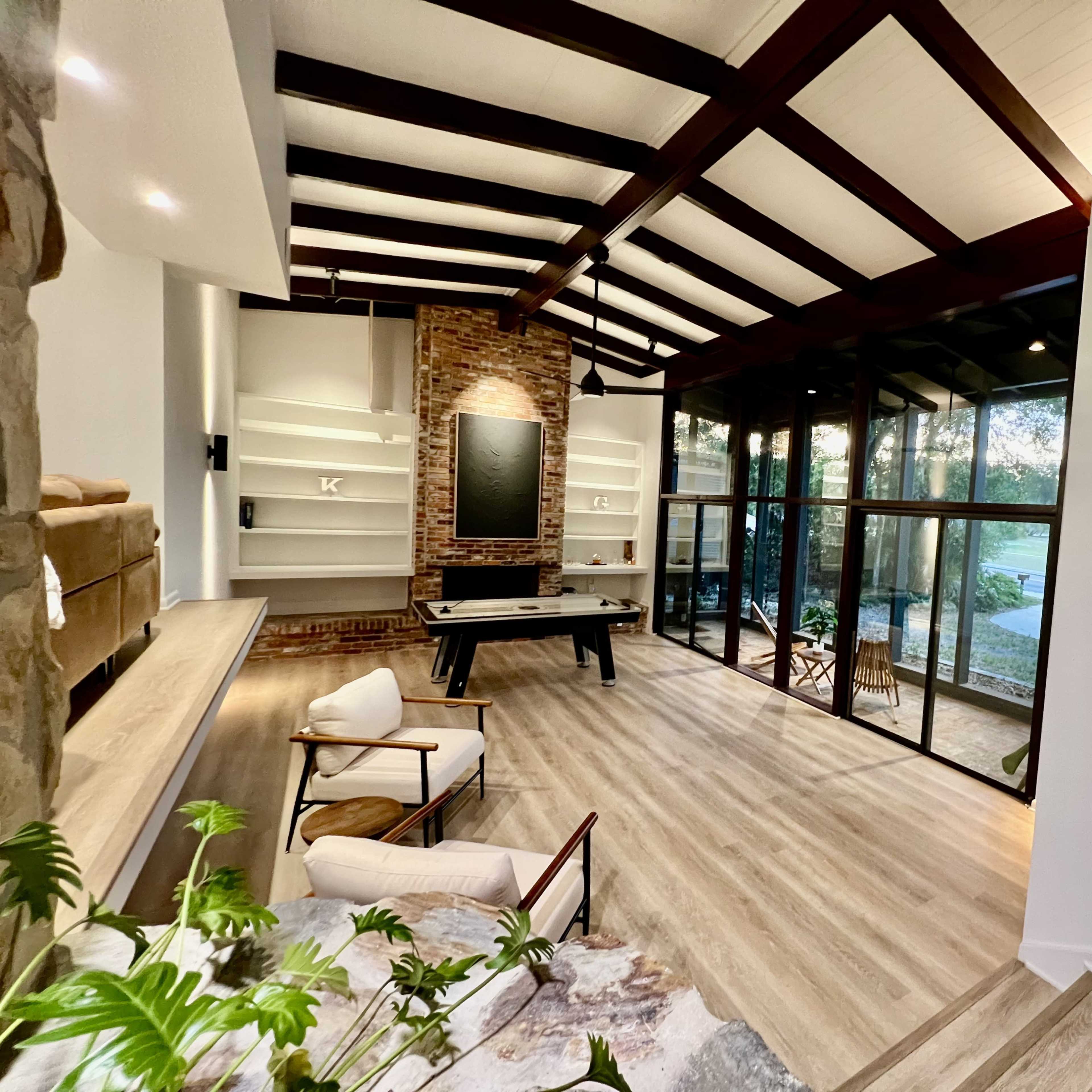 A spacious living area with high, beamed ceilings, large glass windows, and a modern design featuring a pool table and a staircase.