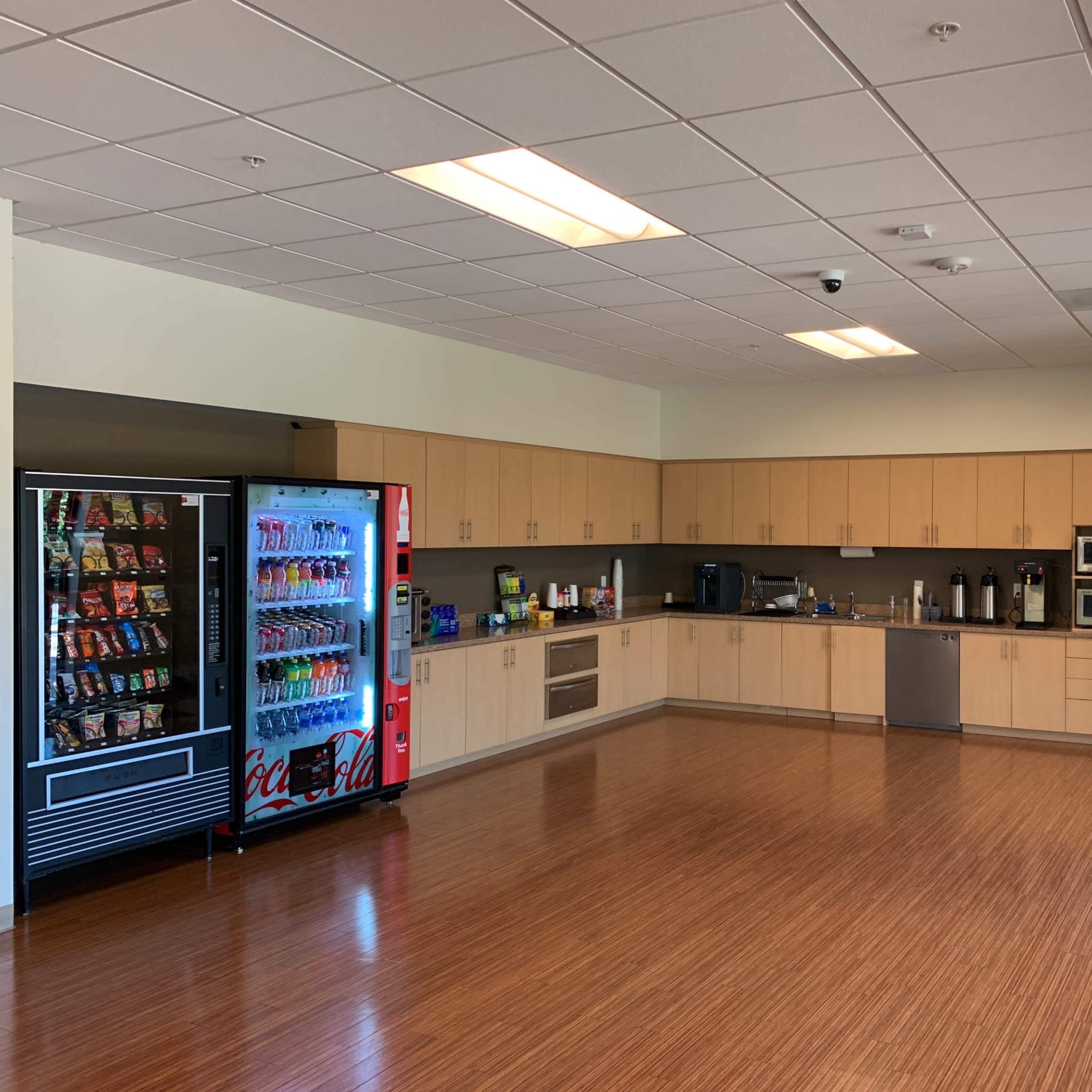 A kitchen area equipped with cabinets, a refrigerator, a microwave, and a beverage vending machine.