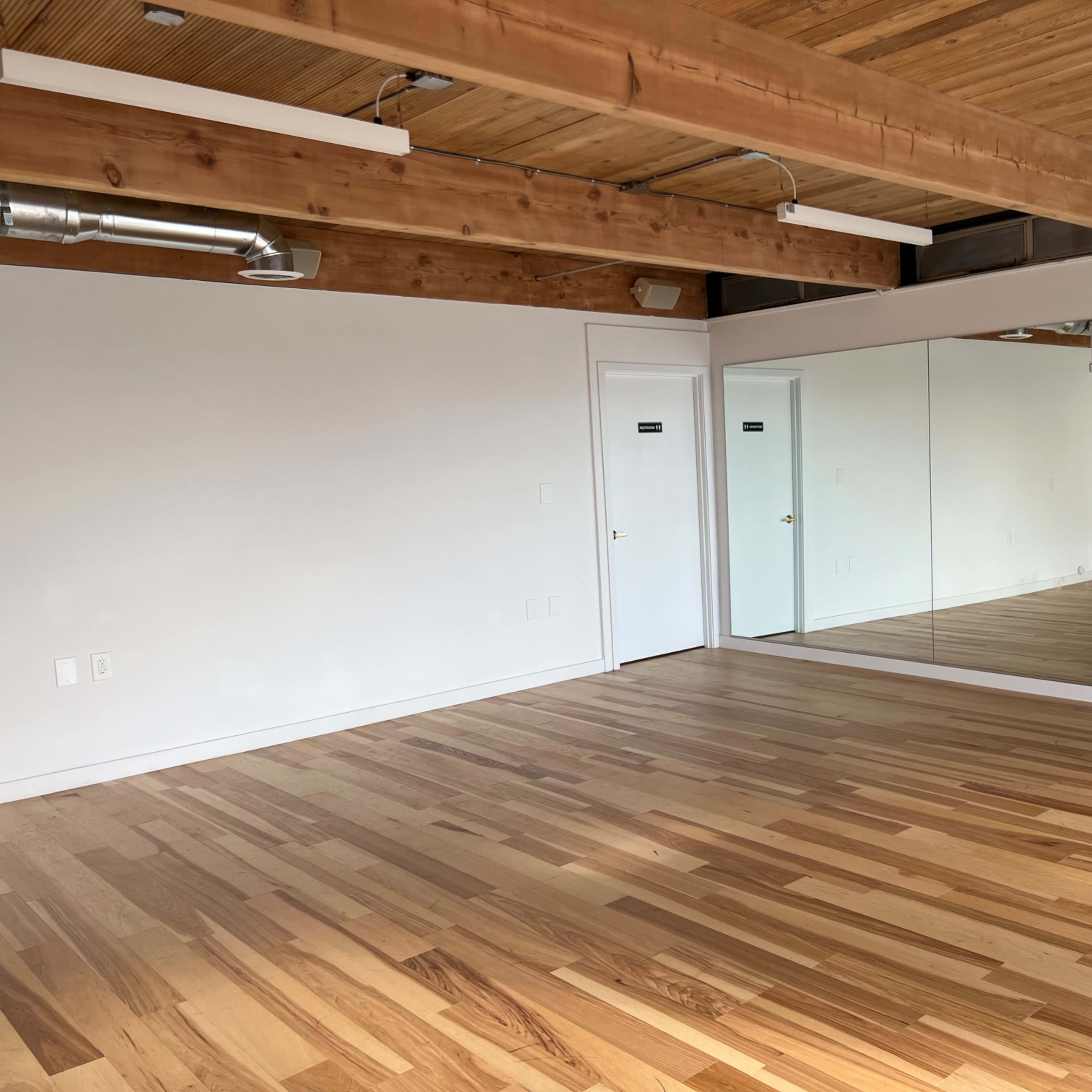 A spacious room with a hardwood floor, exposed wooden beams, and a large mirror on one wall.