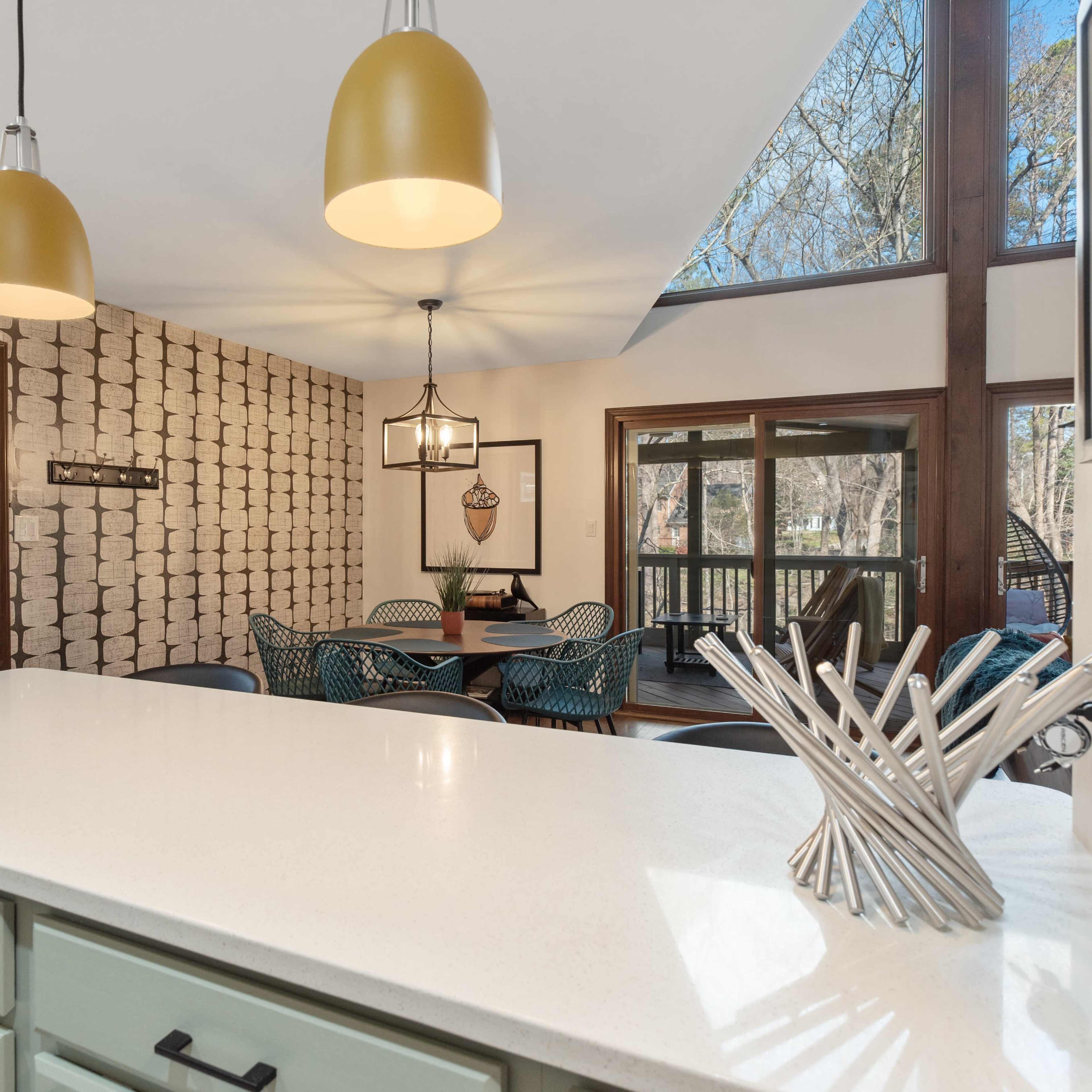 A kitchen area features a white countertop, green cabinets, and pendant lights, with a dining space visible through large windows that overlook an outdoor deck.