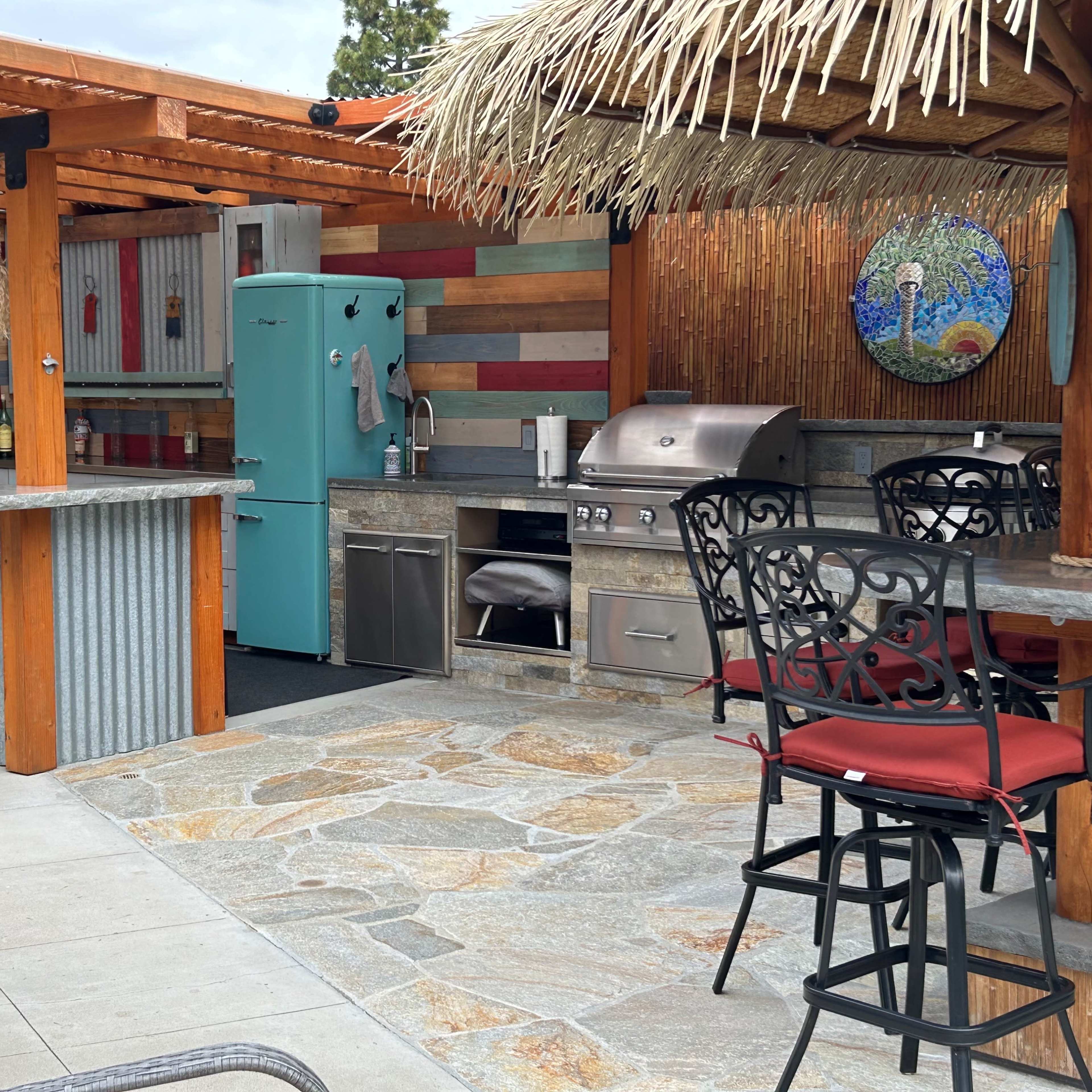 The outdoor kitchen features a stone-paved area with a stainless steel grill, turquoise refrigerator, and bar seating under a thatched roof.