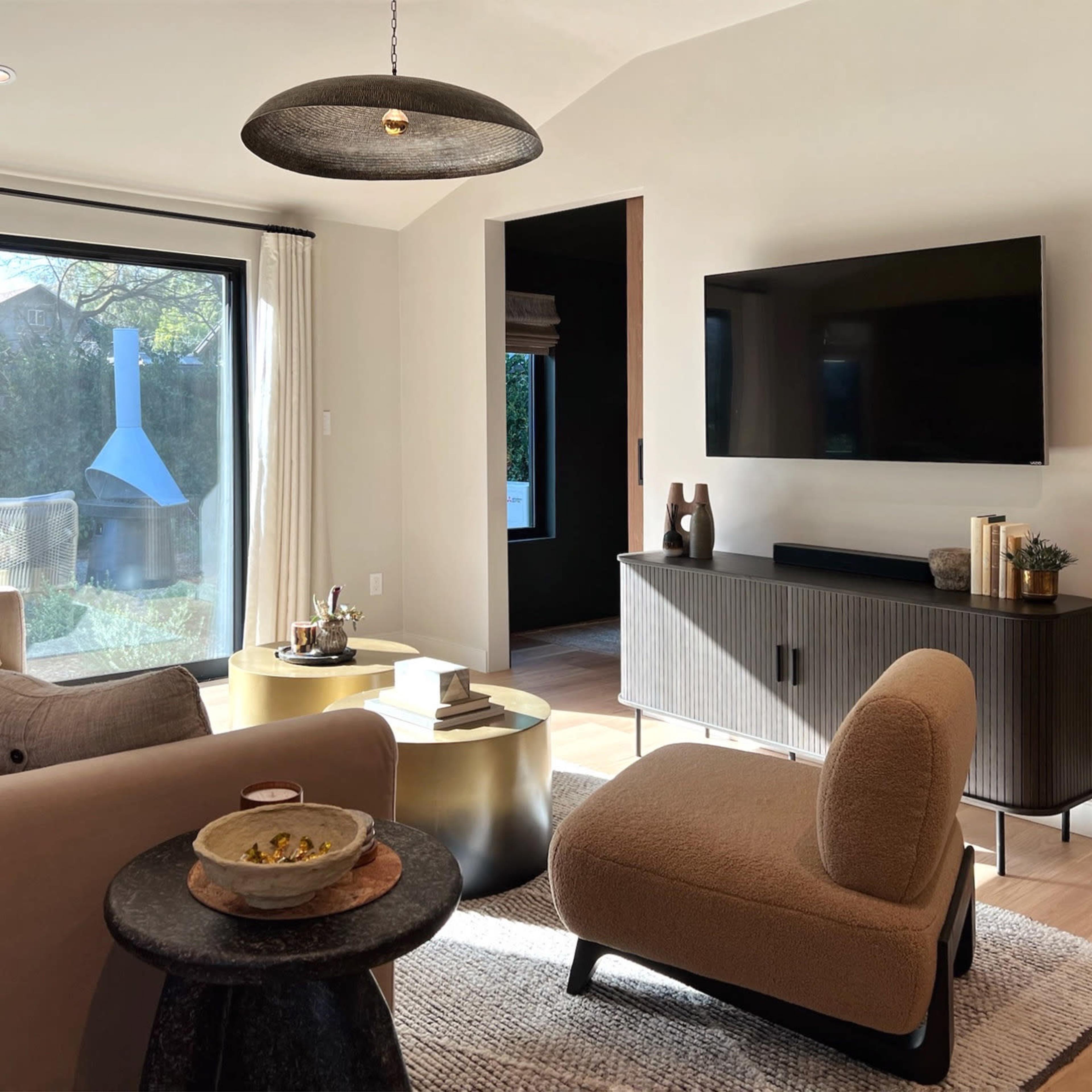 A modern living room with a beige sofa, a round coffee table, a flat-screen TV mounted on the wall, and a cozy armchair, all set against large windows that allow natural light to enter the space.