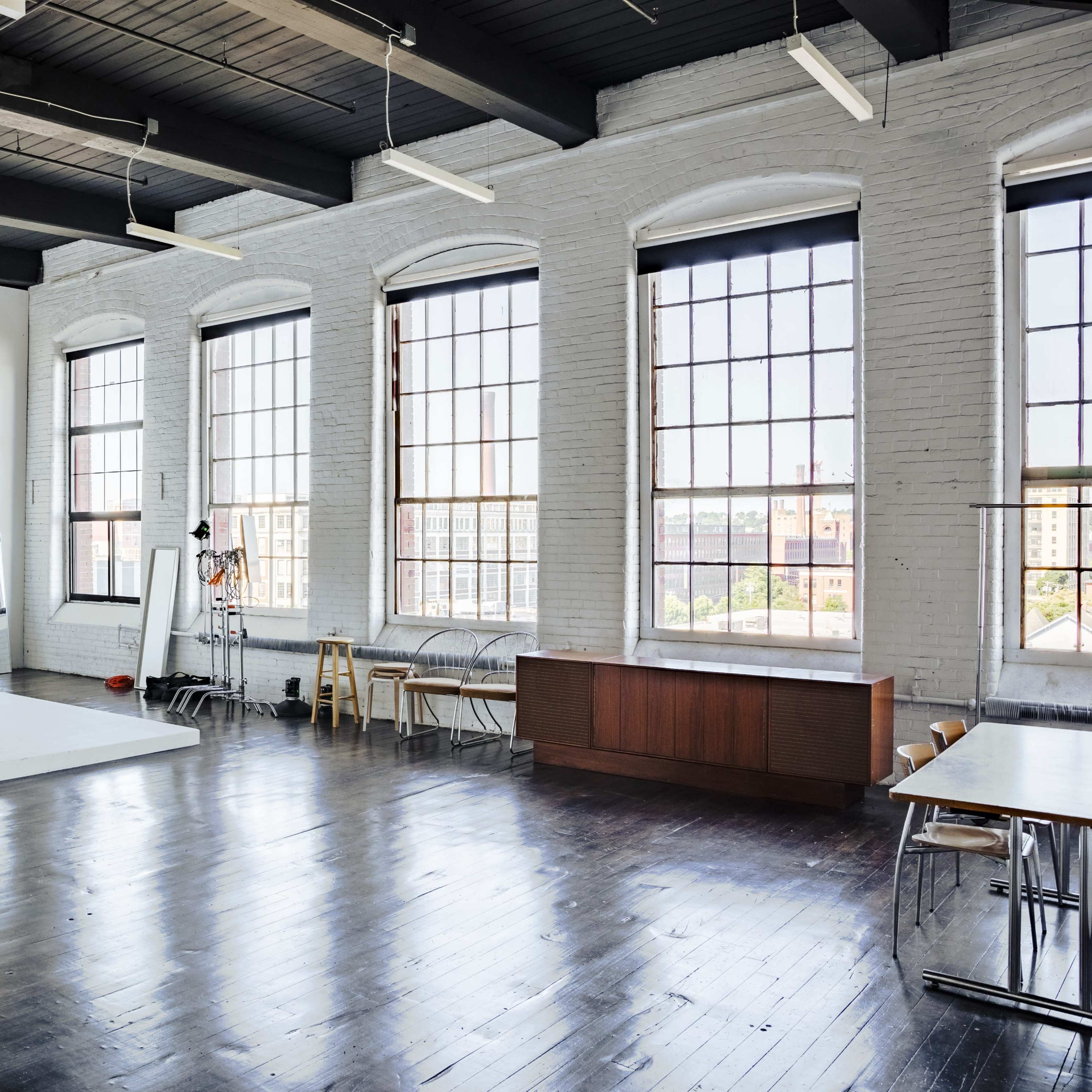 A spacious photography studio with large windows, a plain white backdrop, and several pieces of furniture arranged along the walls.