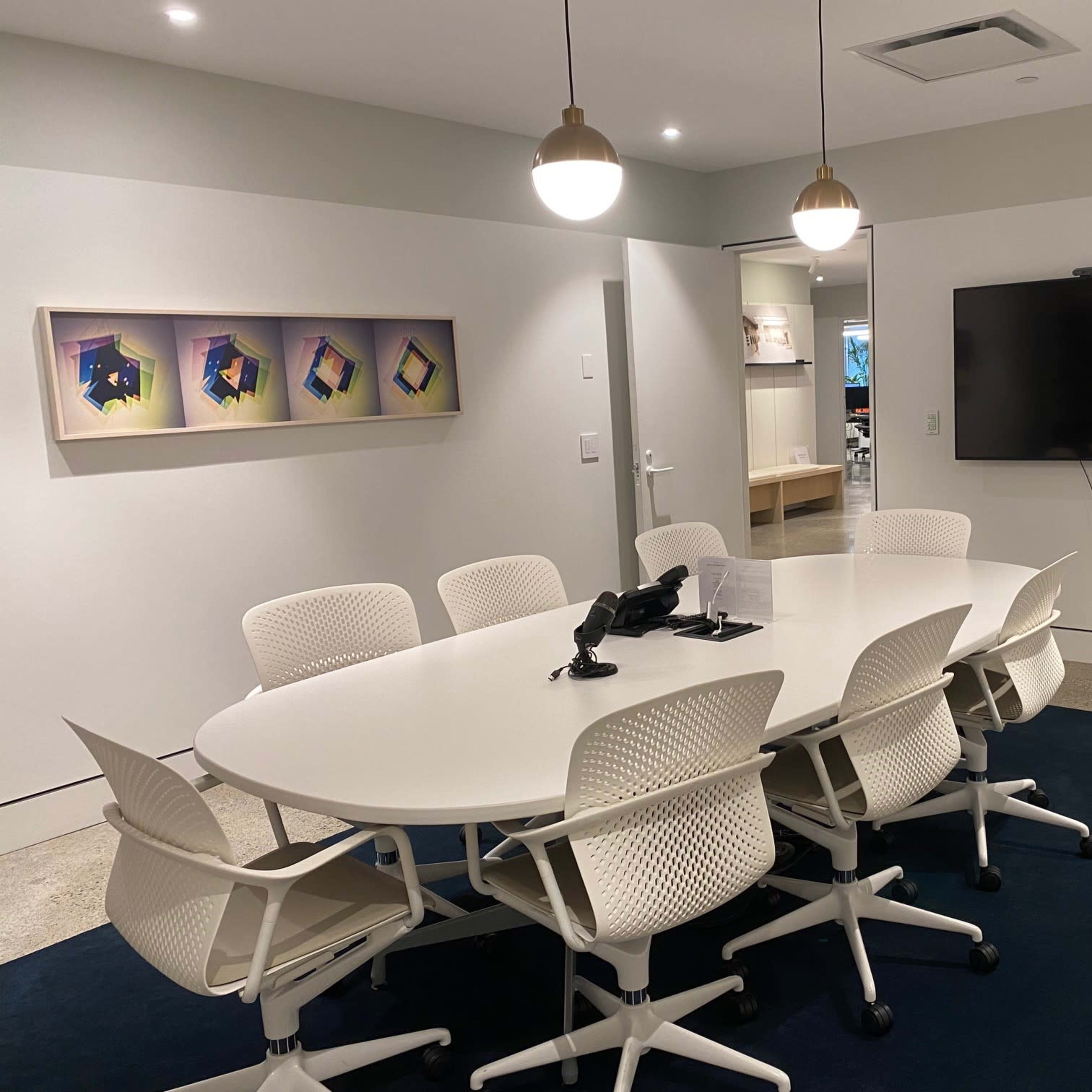 A modern conference room with an oval table surrounded by white chairs, a television mounted on the wall, and a colorful artwork displayed.