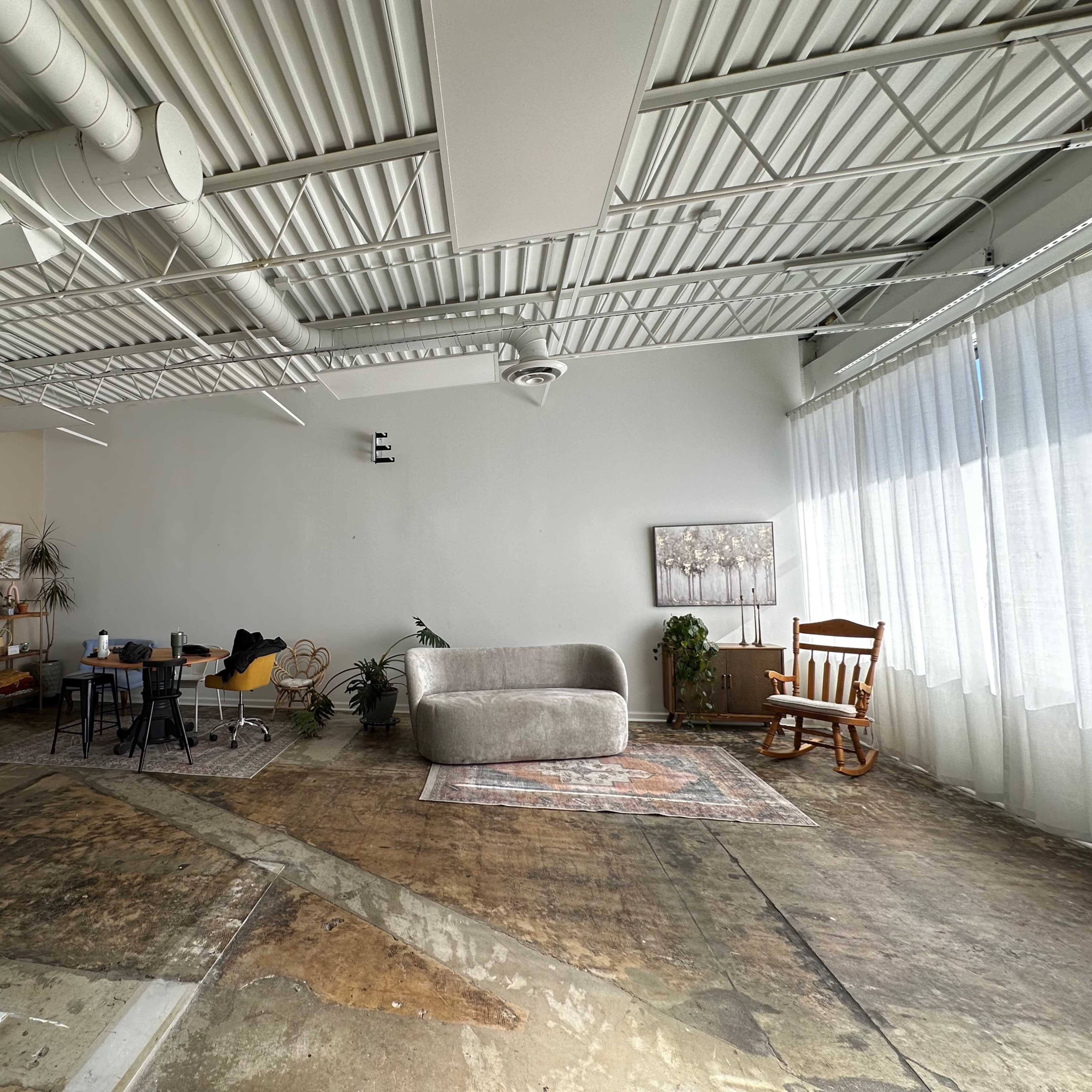 A spacious, industrial-style room with exposed ceilings, a large window, a couch, a dining table with chairs, and a rocking chair, all set against a concrete floor.