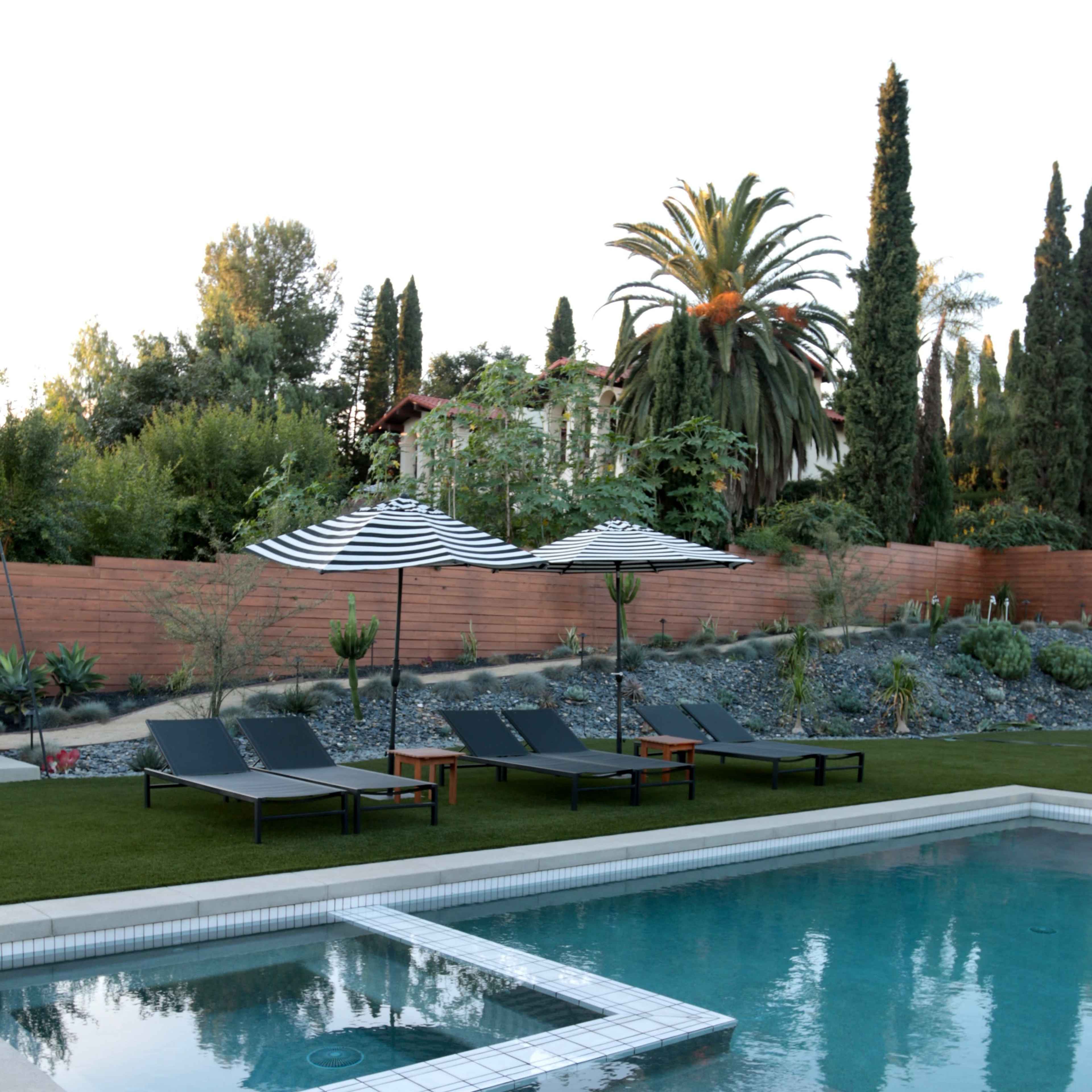 A clear swimming pool surrounded by lounge chairs and umbrellas, with landscaped greenery in the background.
