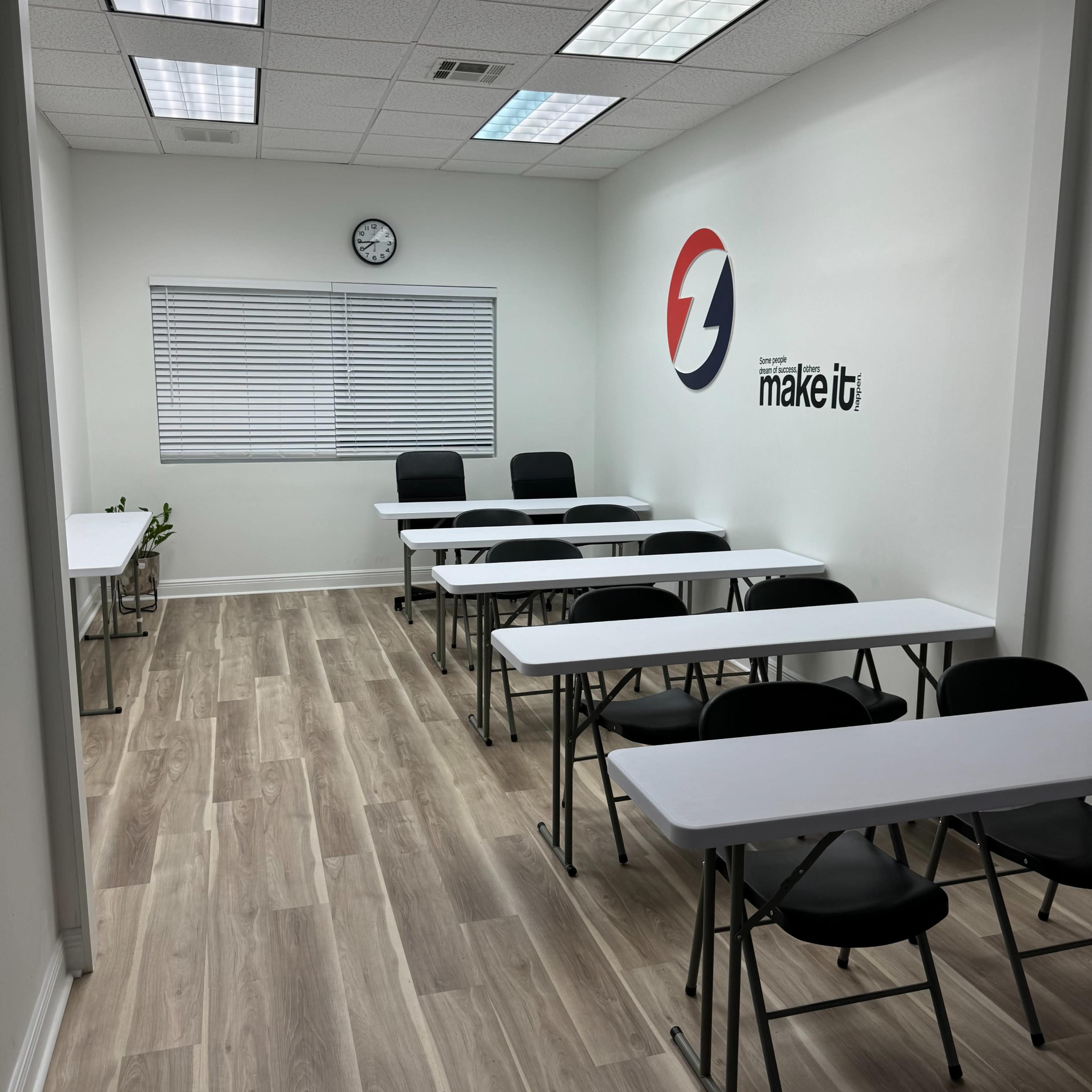 A classroom with several white tables and black chairs arranged neatly, along with a wall clock and a logo on the wall.
