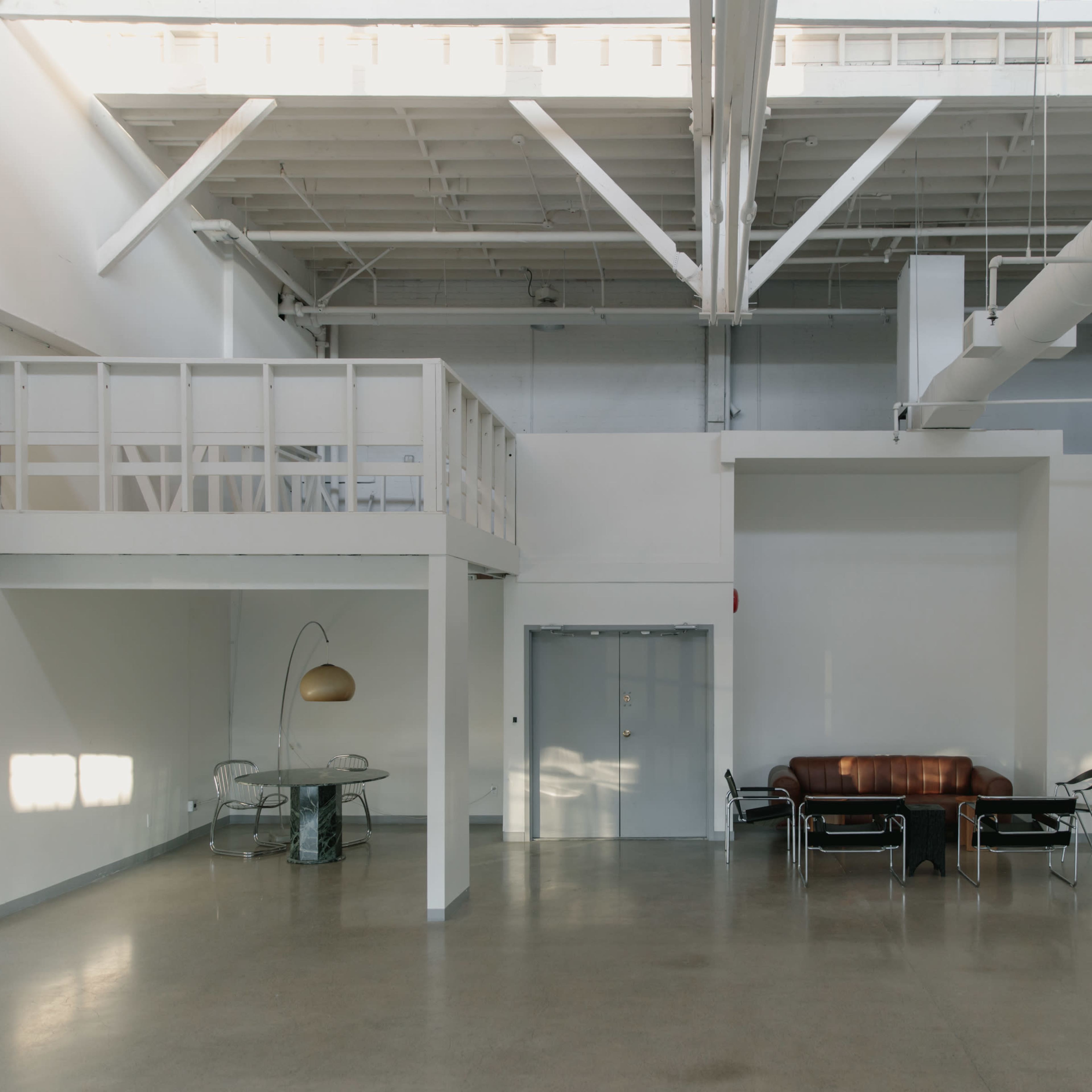 The interior of a spacious, minimalist loft features a mezzanine, a brown leather sofa, a glass dining table with chairs, and large windows allowing natural light to enter.