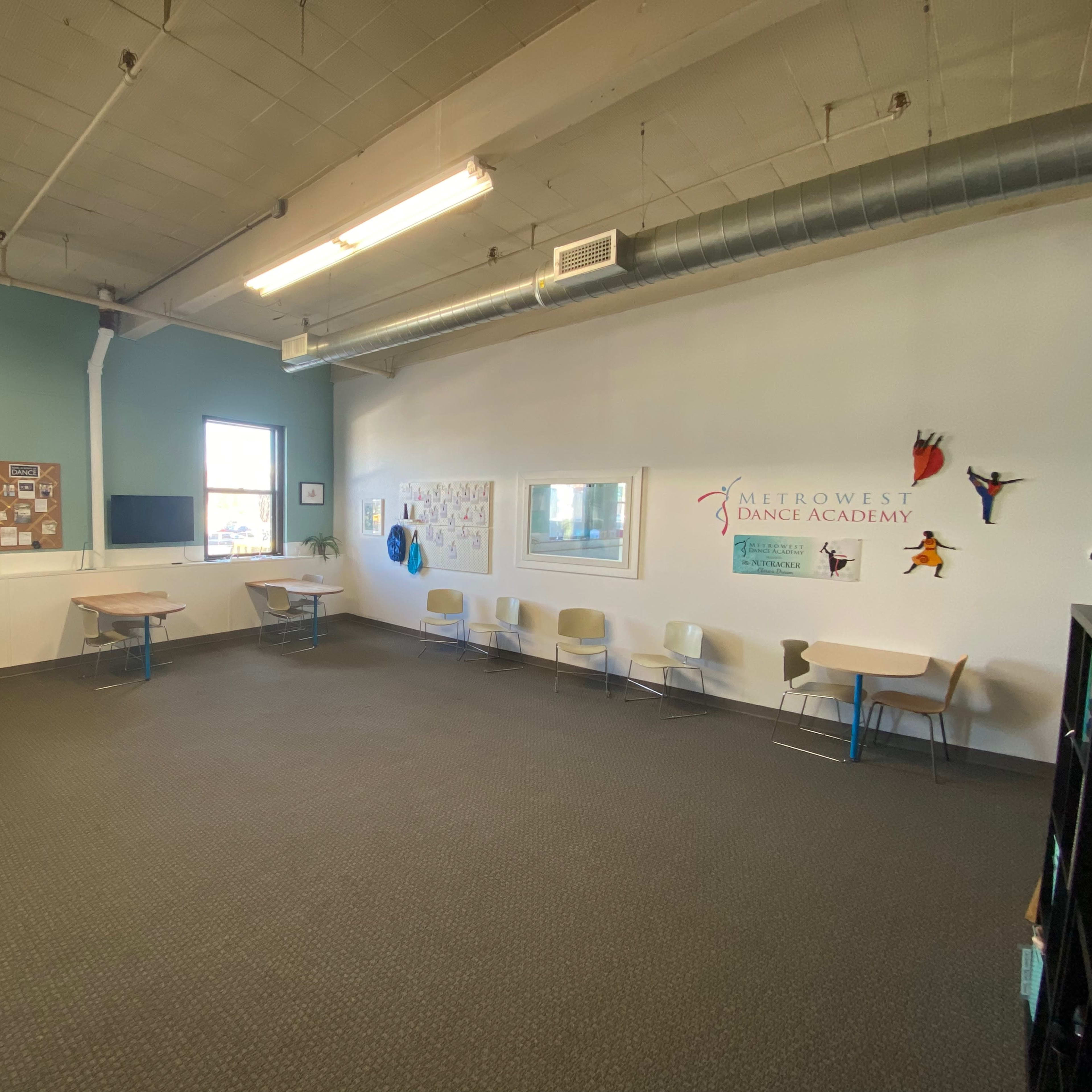 A spacious dance studio with light-colored walls, several tables and chairs arranged along the perimeter, and a large window allowing natural light in.