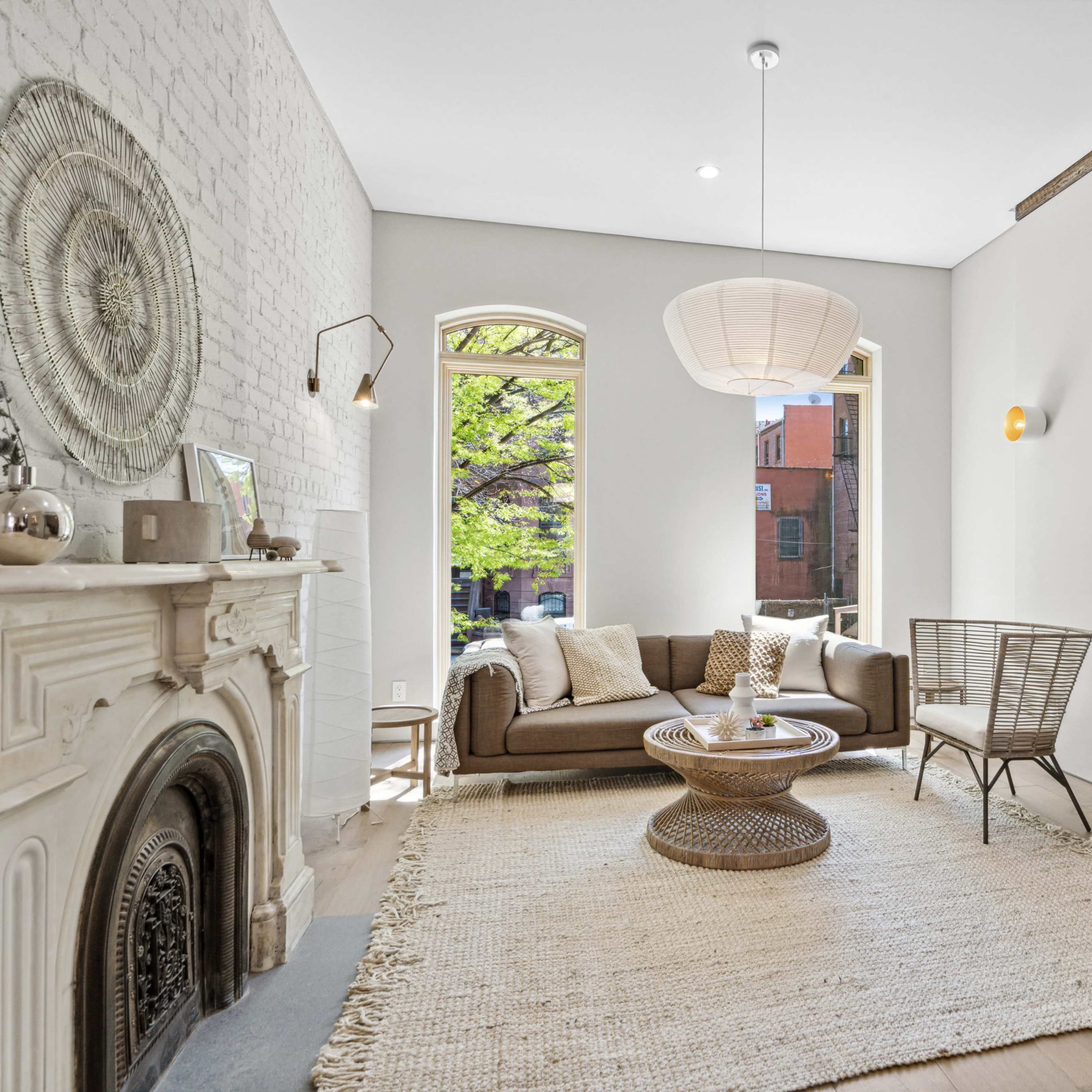 A modern living room with a brown sofa, a round coffee table, a woven rug, and exposed brick walls, along with large windows allowing natural light to enter.