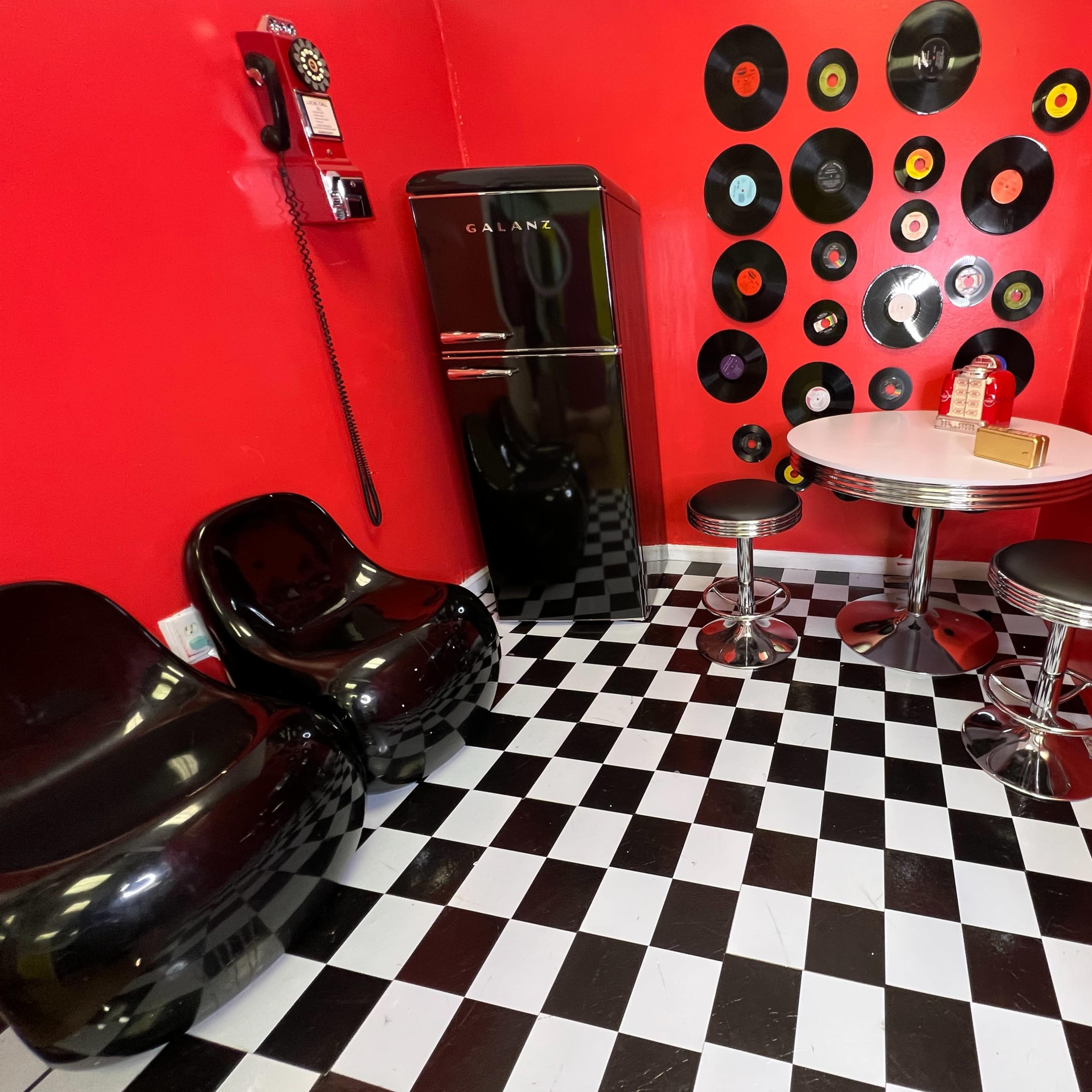 The scene features a small room with red walls adorned with vinyl records, a black refrigerator, two black chairs, and a round table with black and silver stools on a black and white checkered floor.
