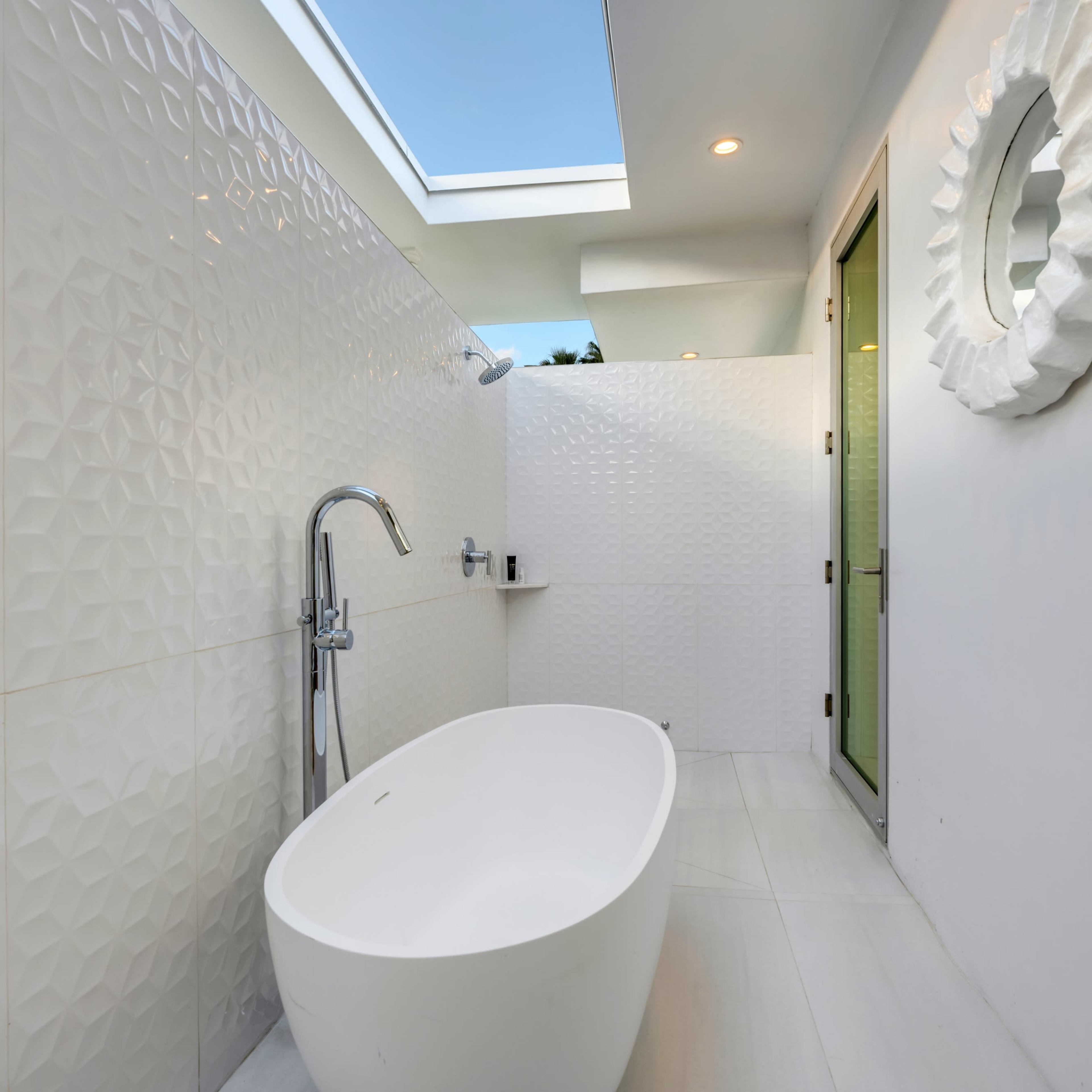 A modern bathroom featuring a freestanding white bathtub, patterned white tiles, and a green door leading outside.