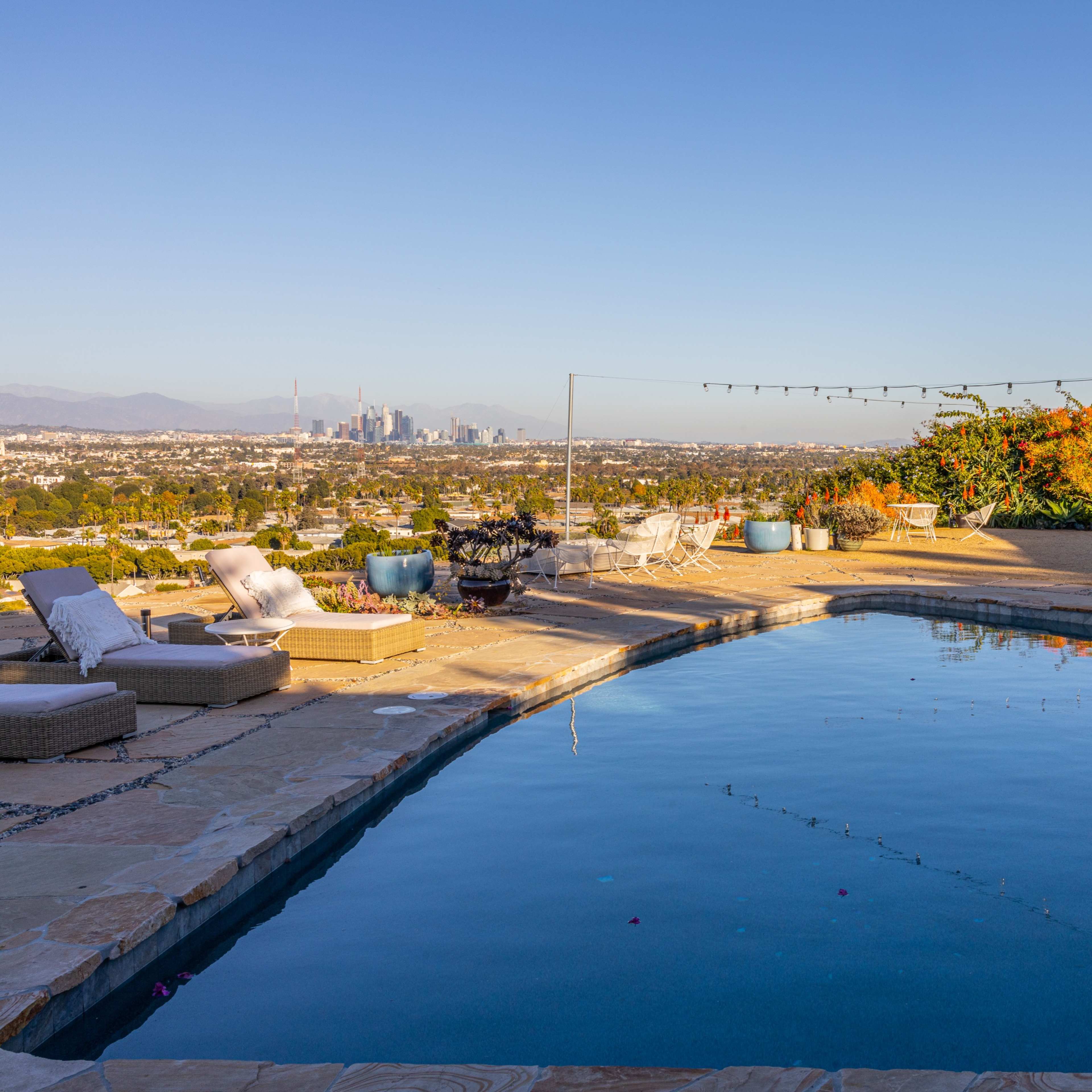 A luxurious swimming pool surrounded by lounge chairs and gardens, overlooking a city skyline.