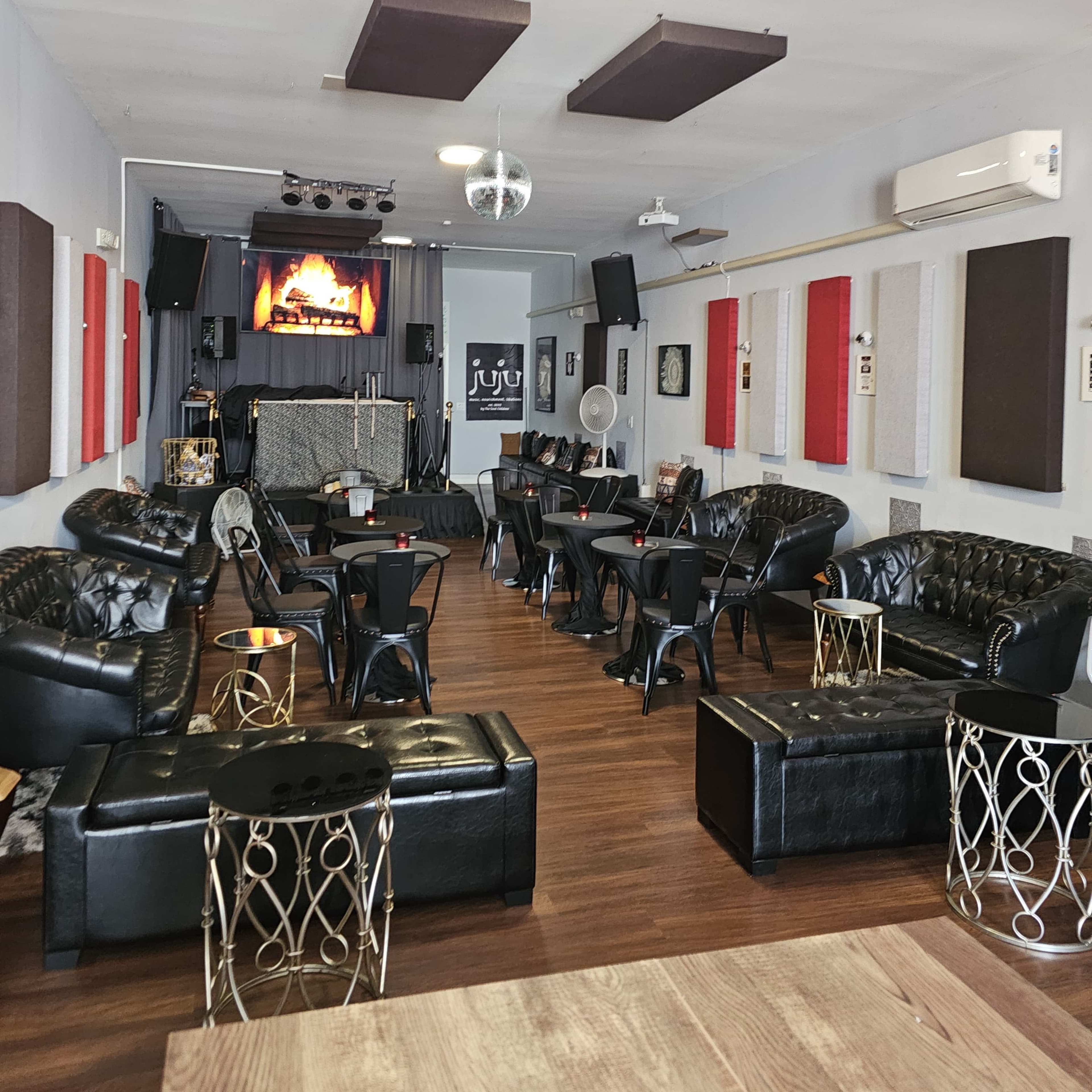 A modern lounge with black leather seating arranged around small tables and a stage at the front featuring a large screen.