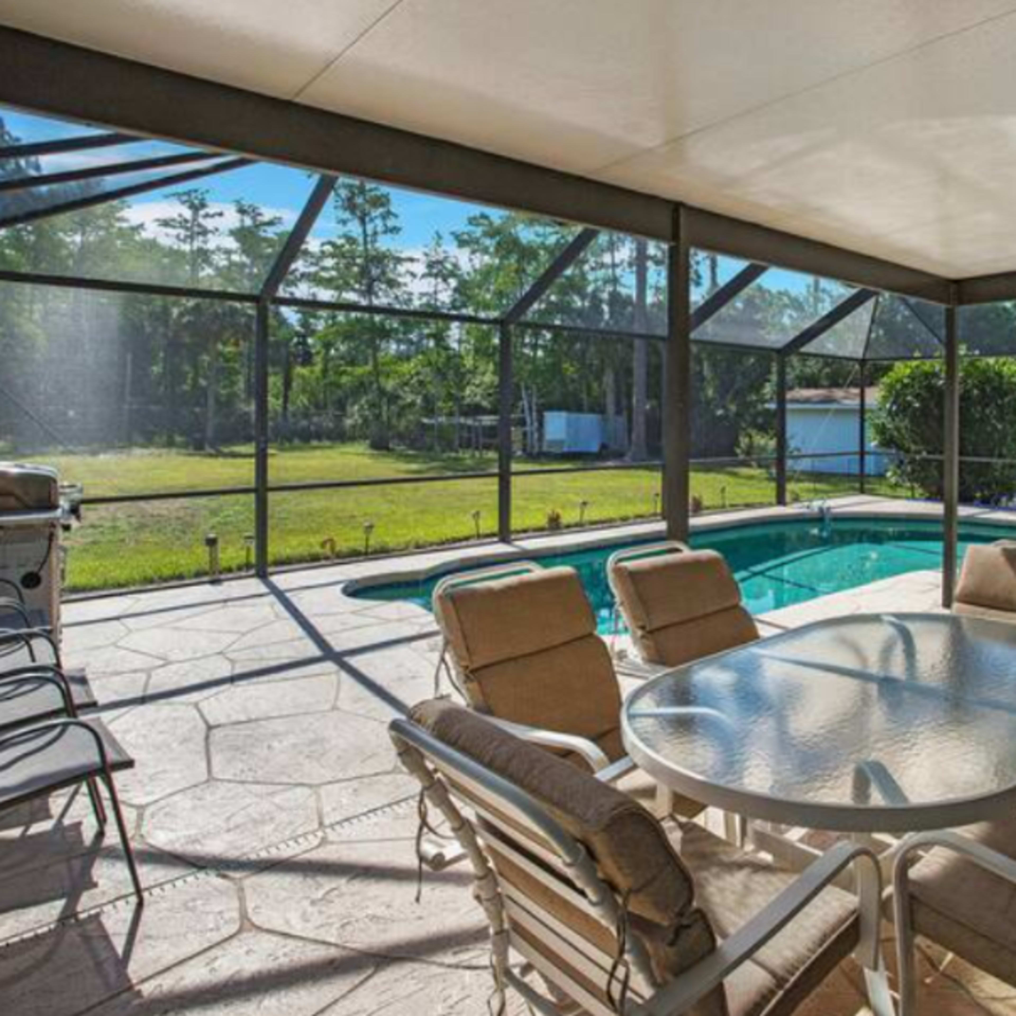 A screened-in patio with a table and chairs overlooking a swimming pool and green backyard.