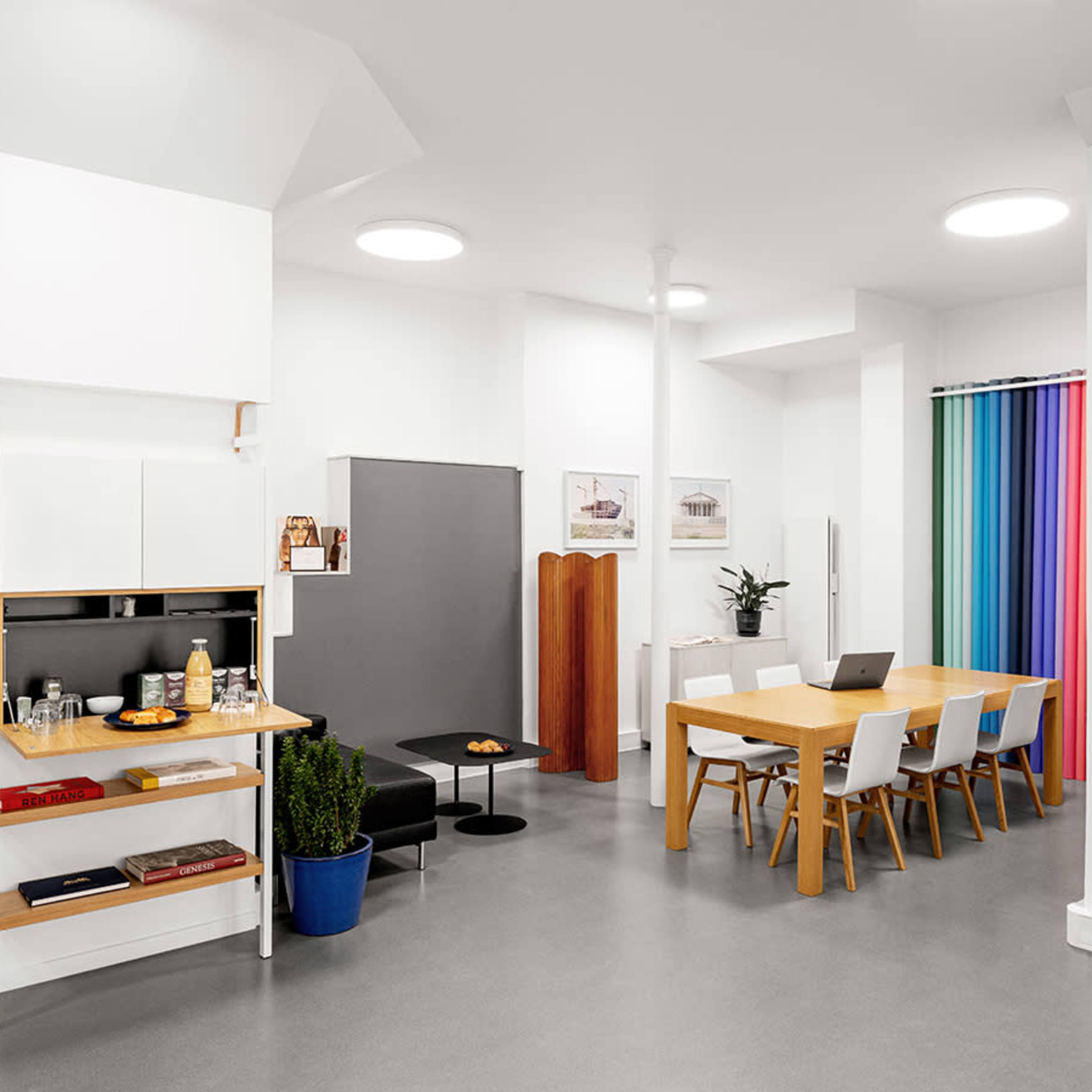 A modern, minimalist kitchen and dining area with a wooden table, white chairs, and a colorful curtain in the background.