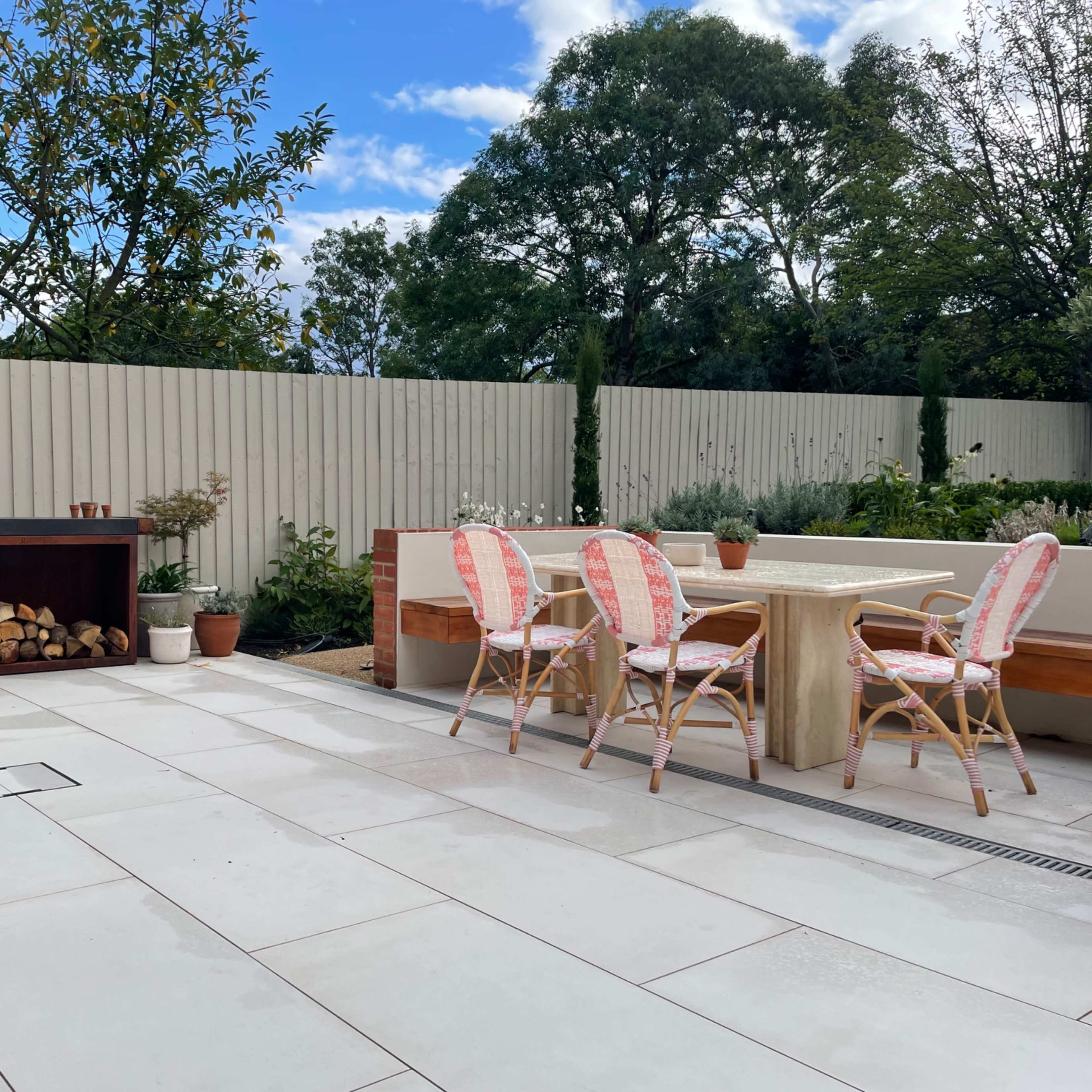 The scene features a patio area with a wooden table and chairs, a firewood storage unit, and potted plants, surrounded by a fence and green trees under a partly cloudy sky.